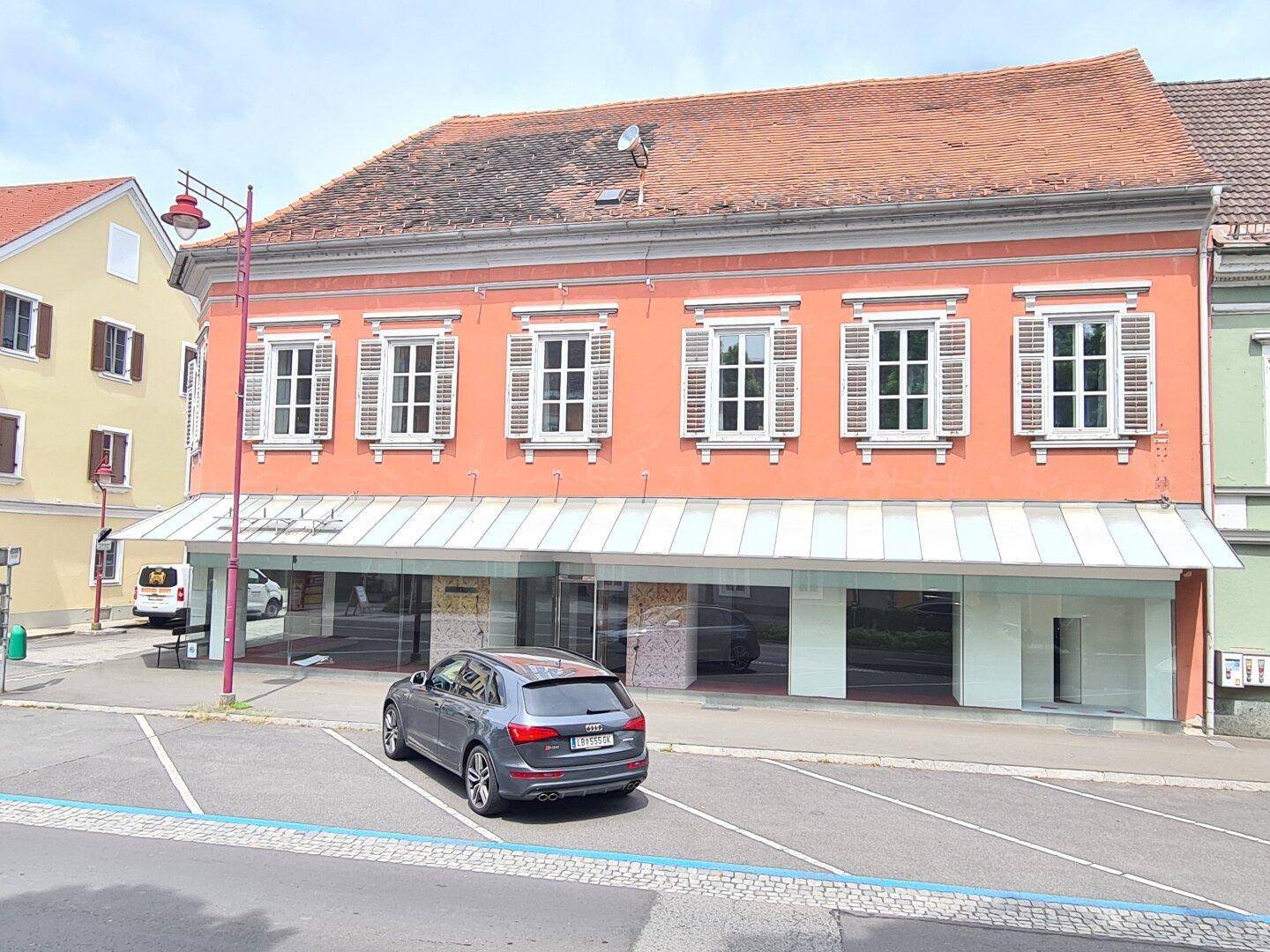 Ansicht des historischen Gebäudes mit rosafarbener Fassade, Schaufenstern und einem geparkten Auto davor.