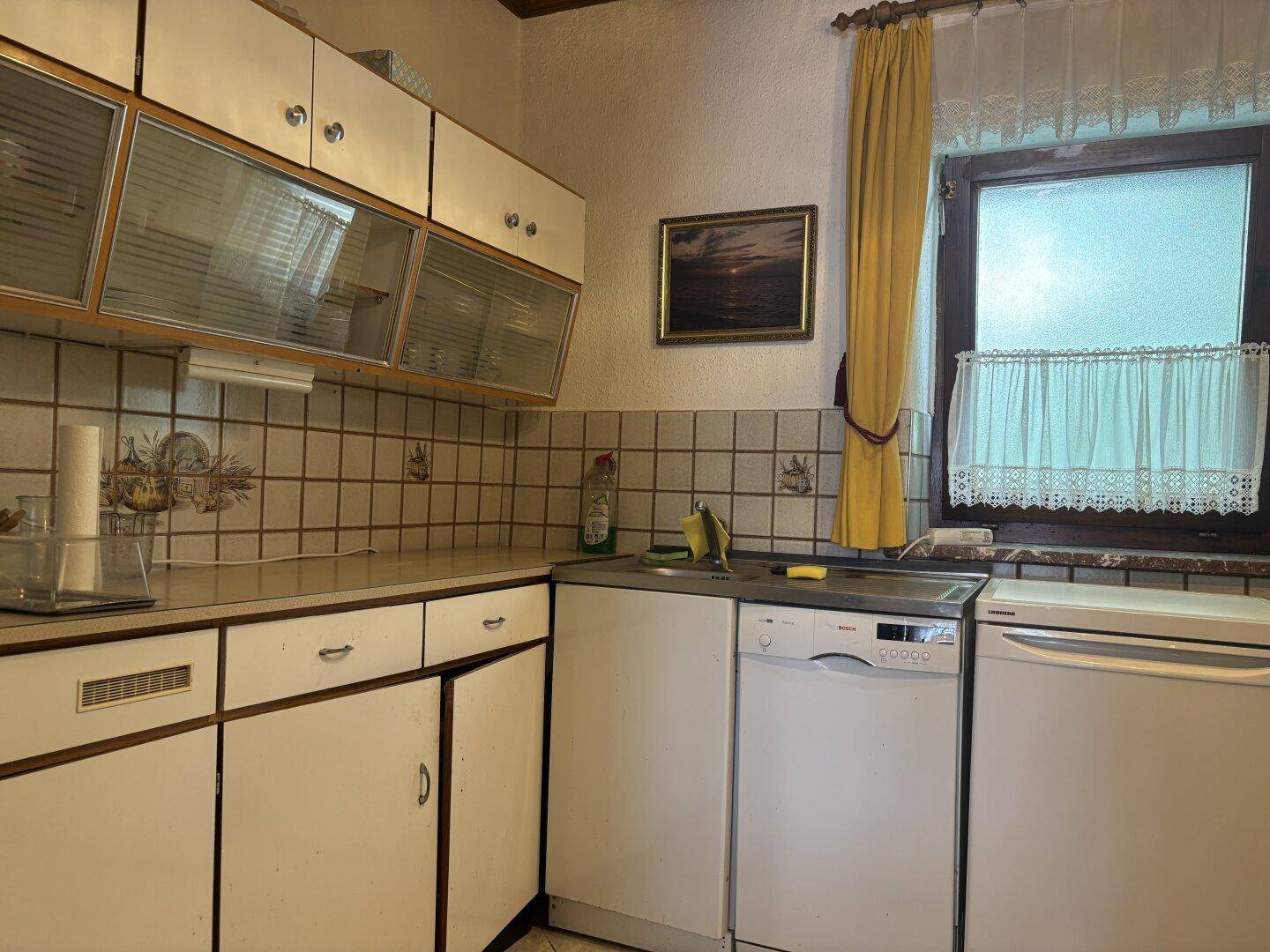 Einfache Küche mit weißen Schränken, Spülmaschine und Kühlschrank neben dem Fenster.