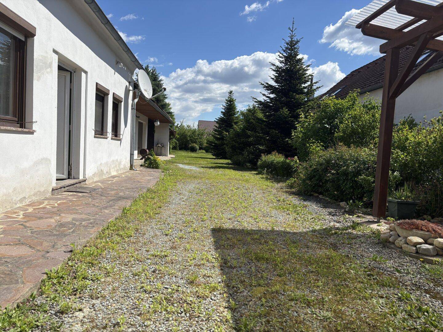 Gepflasterter Weg entlang der Hausseite mit Sträuchern und Bäumen im Hintergrund.