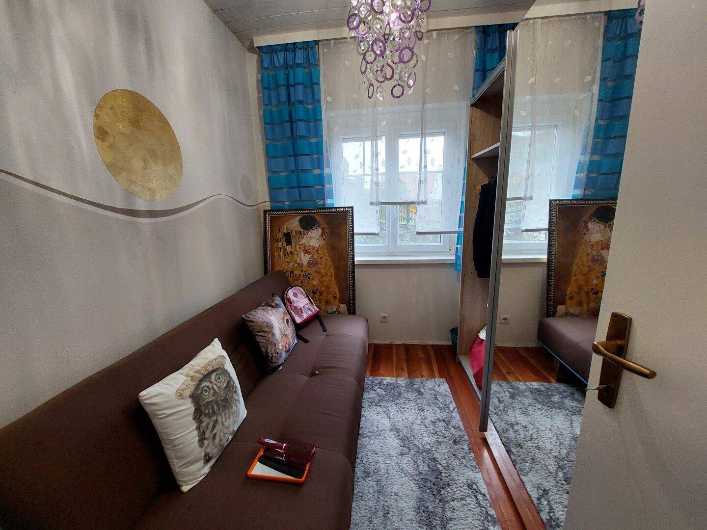 Helles Zimmer mit Schlafsofa, Spiegelschrank und farbigen Vorhängen.