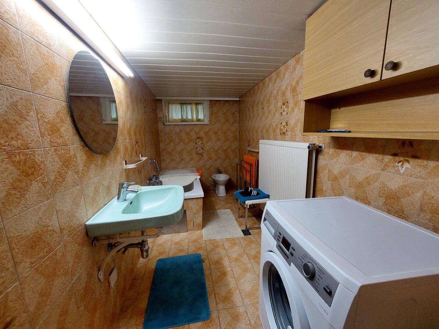 Funktionales Badezimmer mit Badewanne, Bidet, Waschbecken und Waschmaschine in braunen Fliesen.