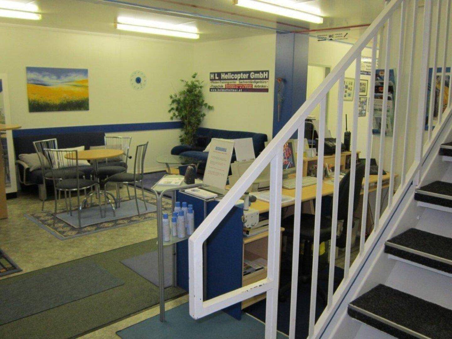 Empfangsbereich mit Schreibtisch, Sitzgelegenheiten und einer Treppe, die in ein Obergeschoss führt.