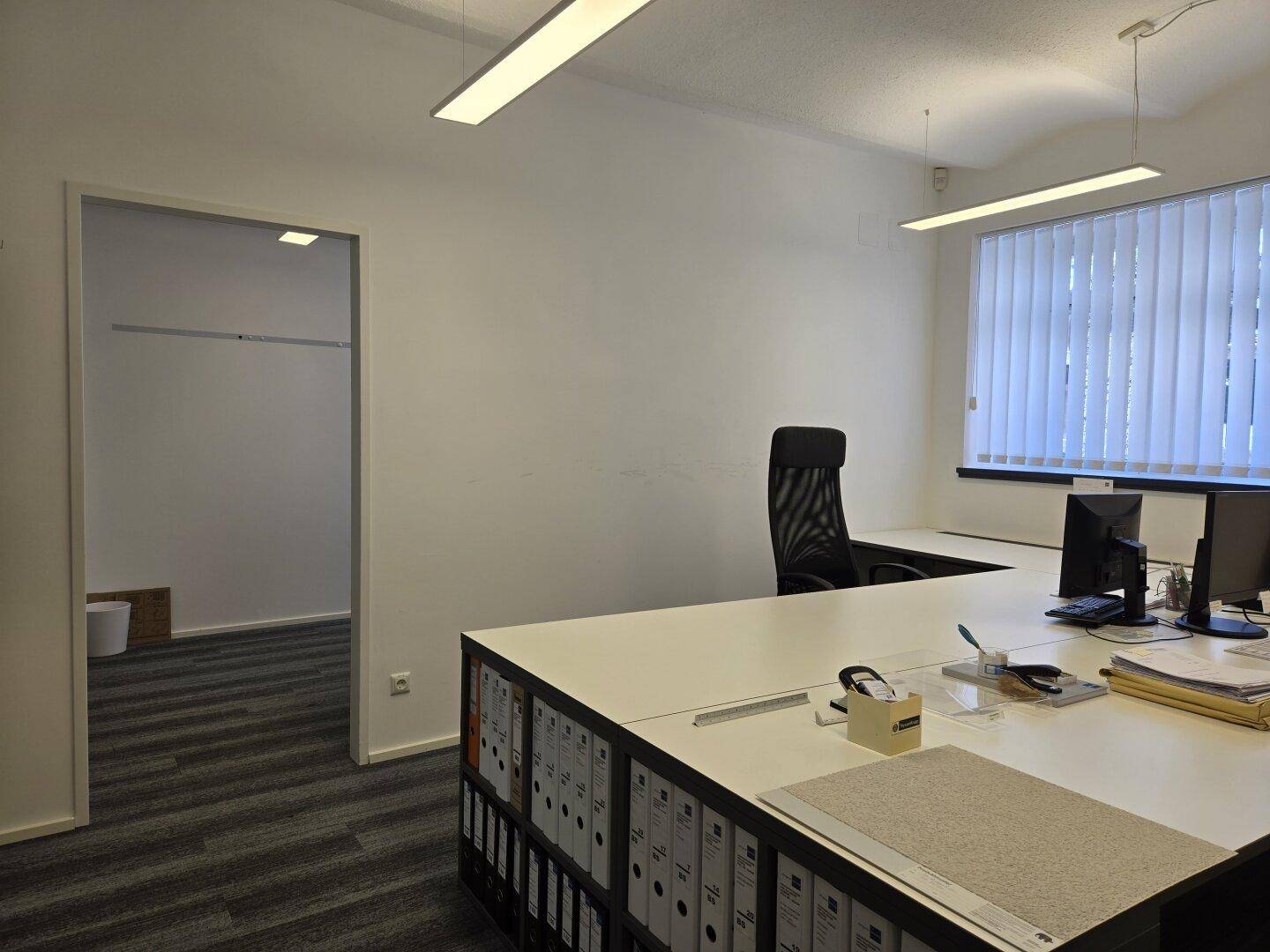 Einzelbüro mit Schreibtisch, Bürostuhl und Fenster mit Jalousien.