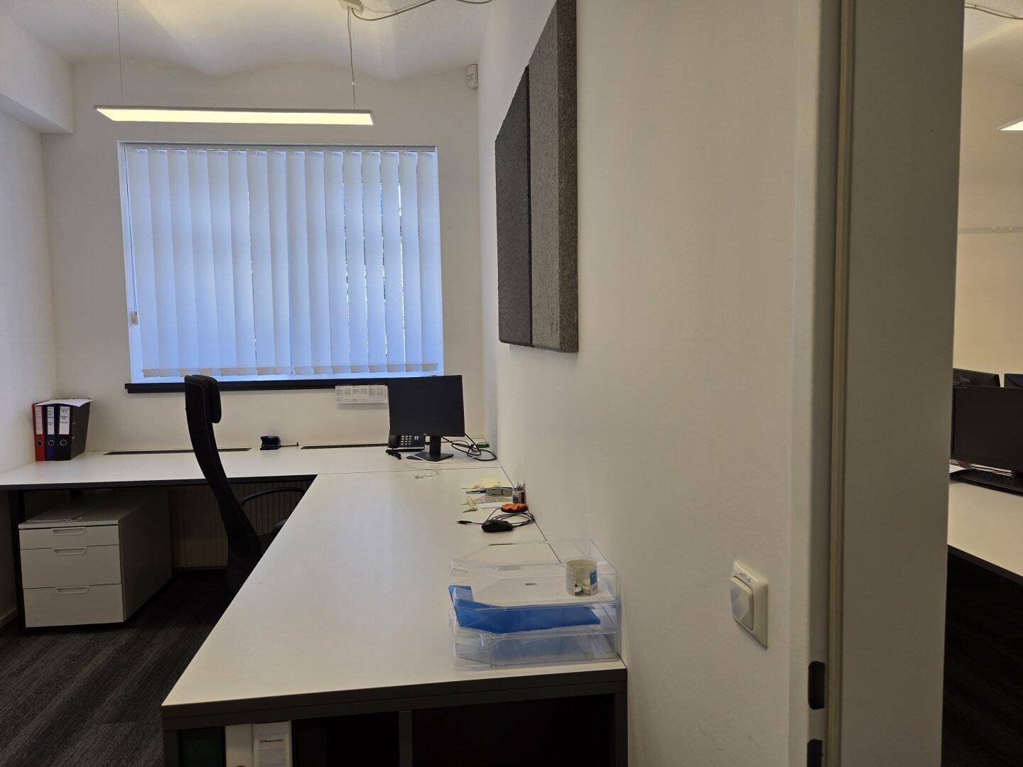 Einzelner Büroarbeitsplatz mit Schreibtisch, Bürostuhl und Fenster mit Jalousien.