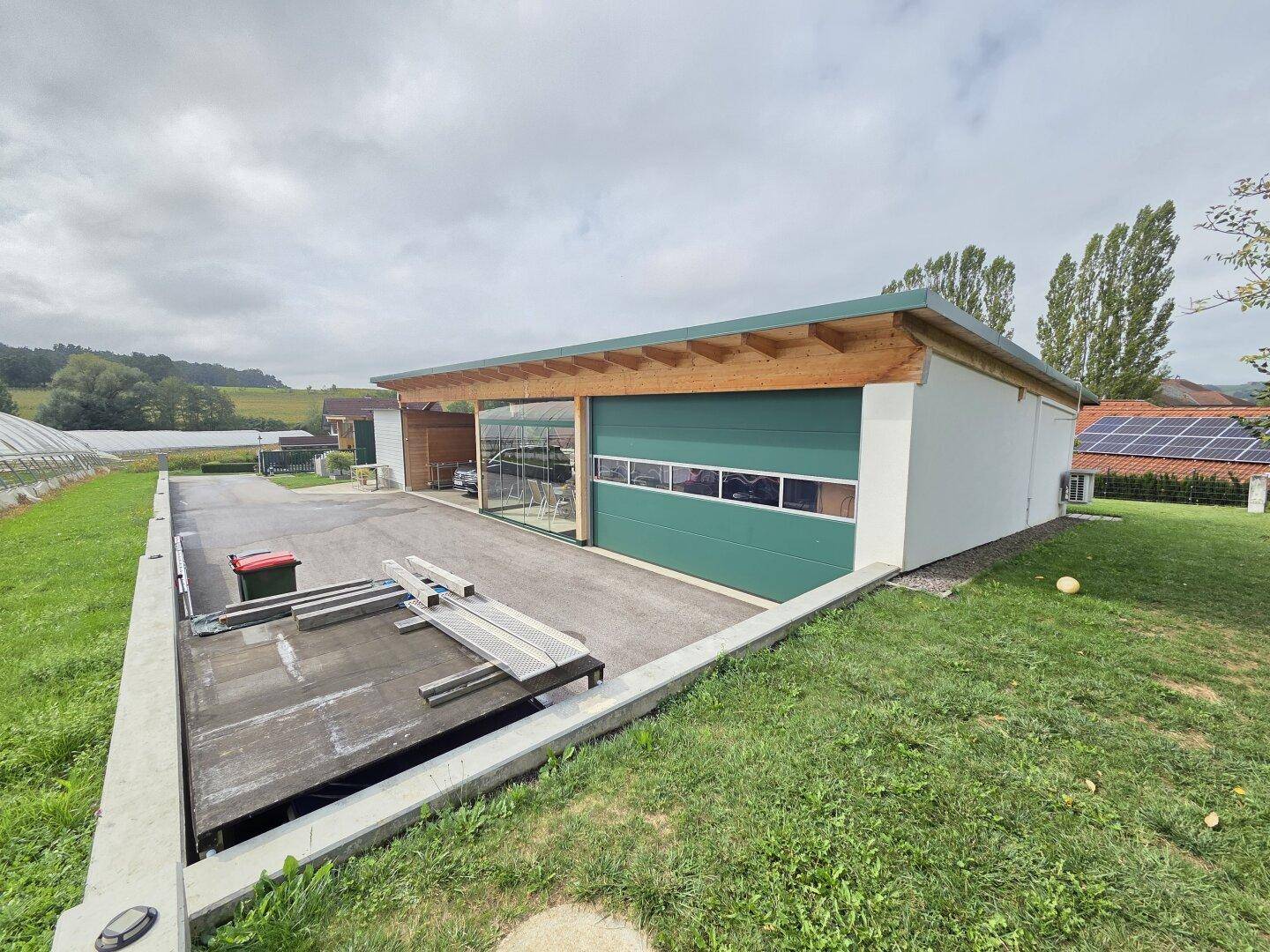 Außenansicht des Gebäudes mit grüner Garagentür, verglastem Bereich und Carport, umgeben von Rasen.