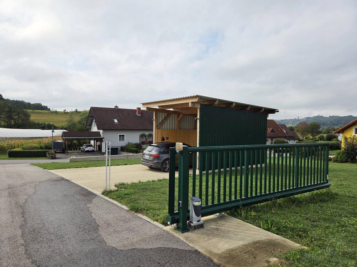 Zufahrt zum Grundstück mit grünem Schiebetor und einem hölzernen Carport, in dem ein Auto parkt.