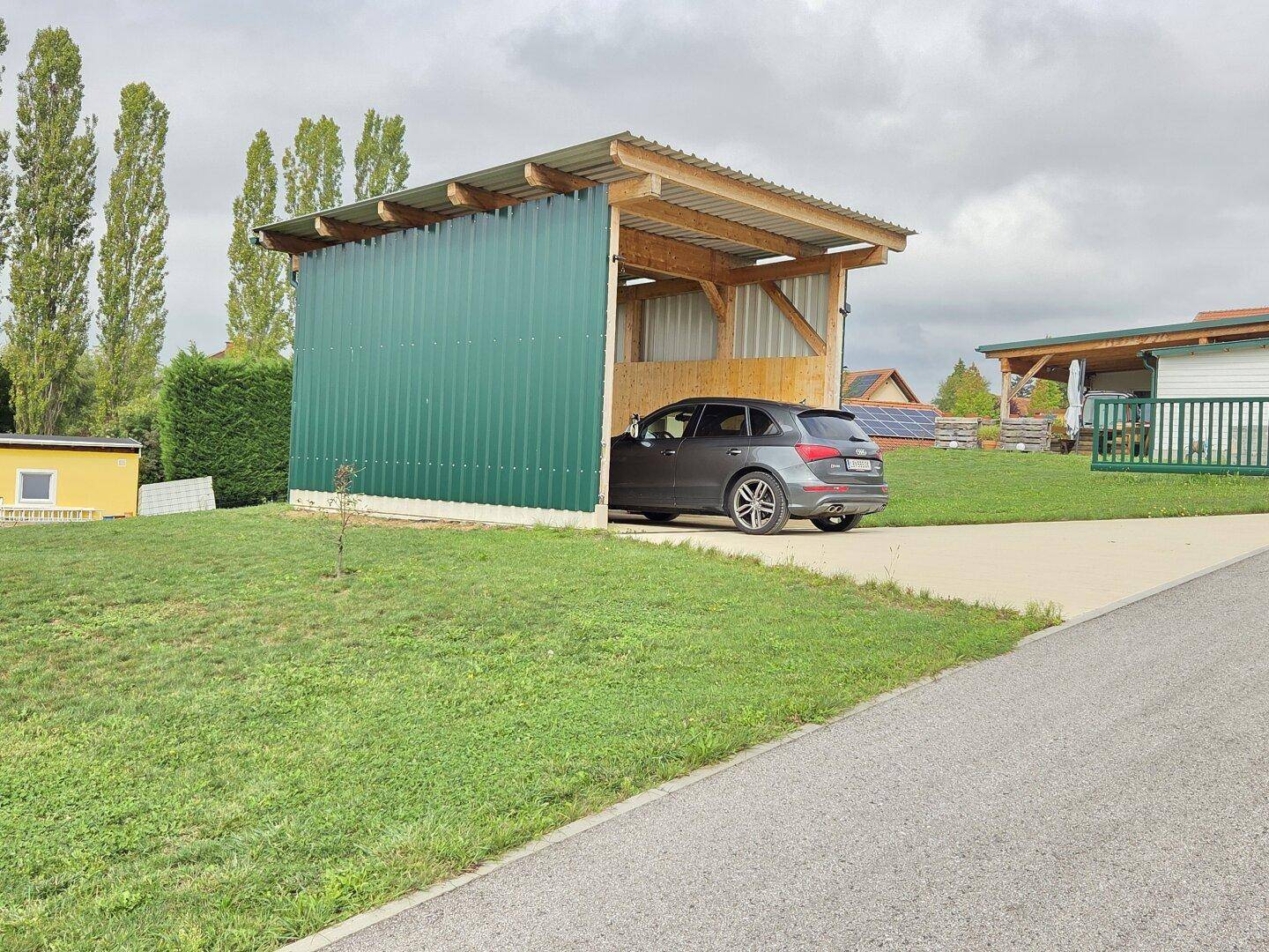 Hölzerner Carport mit grüner Metallverkleidung und einem geparkten Auto, umgeben von einer Rasenfläche.
