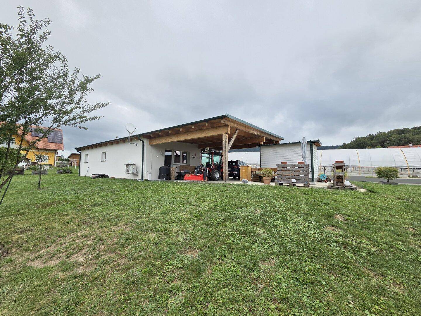 Seitenansicht des weißen Gebäudes mit hölzernem Carport und landwirtschaftlichen Geräten auf dem Rasen.