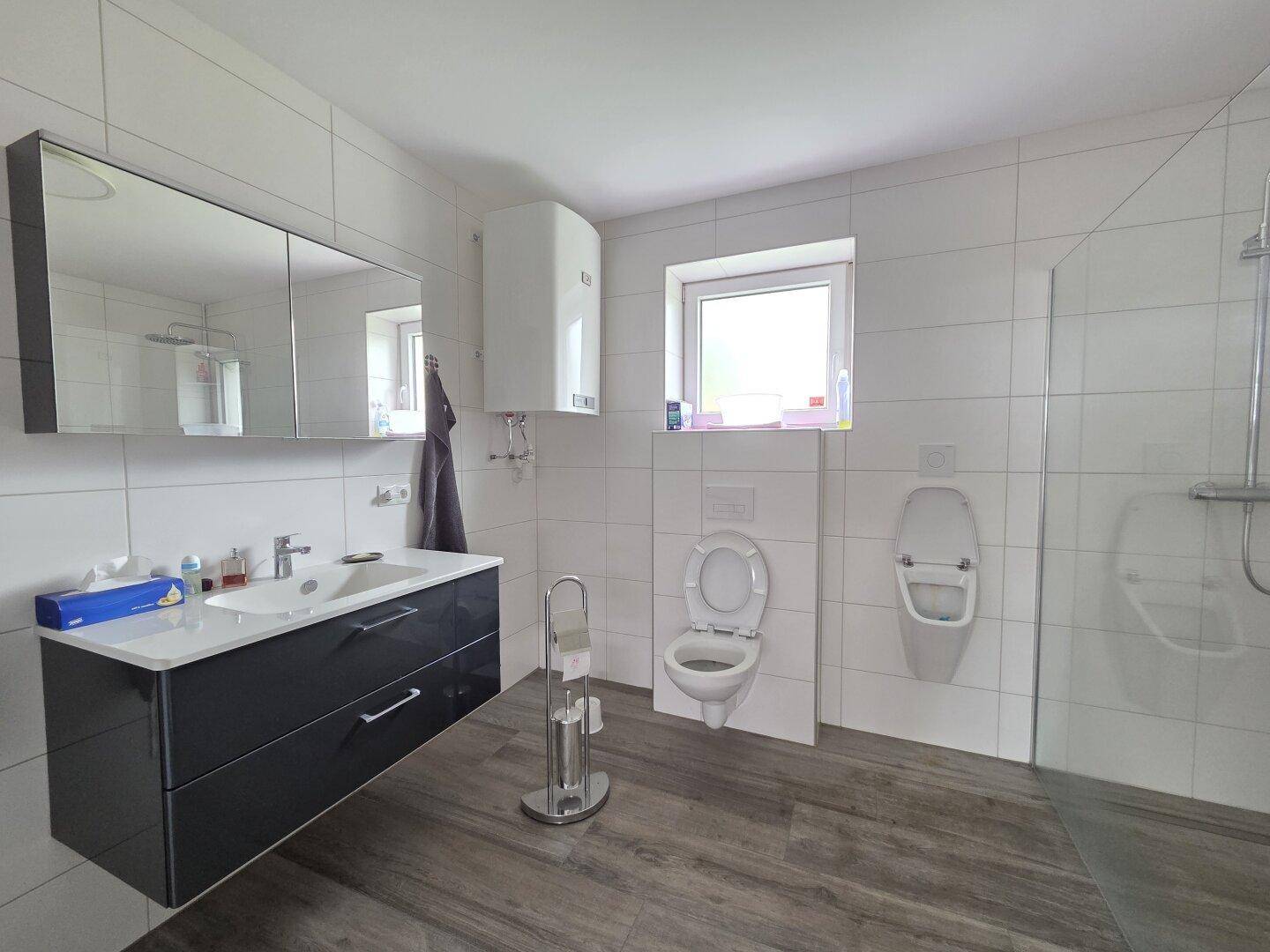 Modernes Badezimmer mit weißen Fliesen, dunklem Waschtisch, Toilette, Urinal und einer begehbaren Dusche.