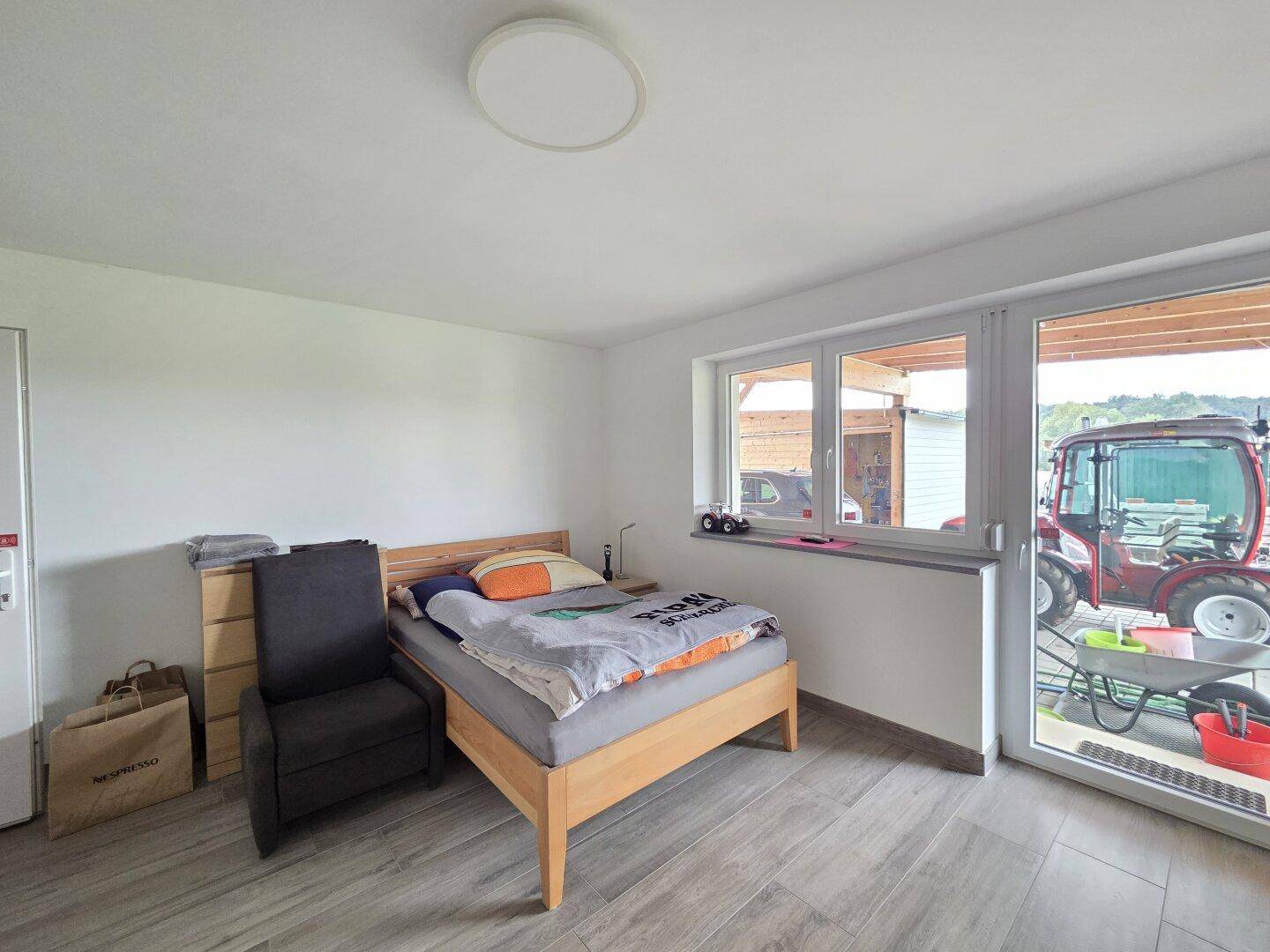 Helles Schlafzimmer mit Holzbett und großem Fenster, das den Blick auf den Außenbereich freigibt.