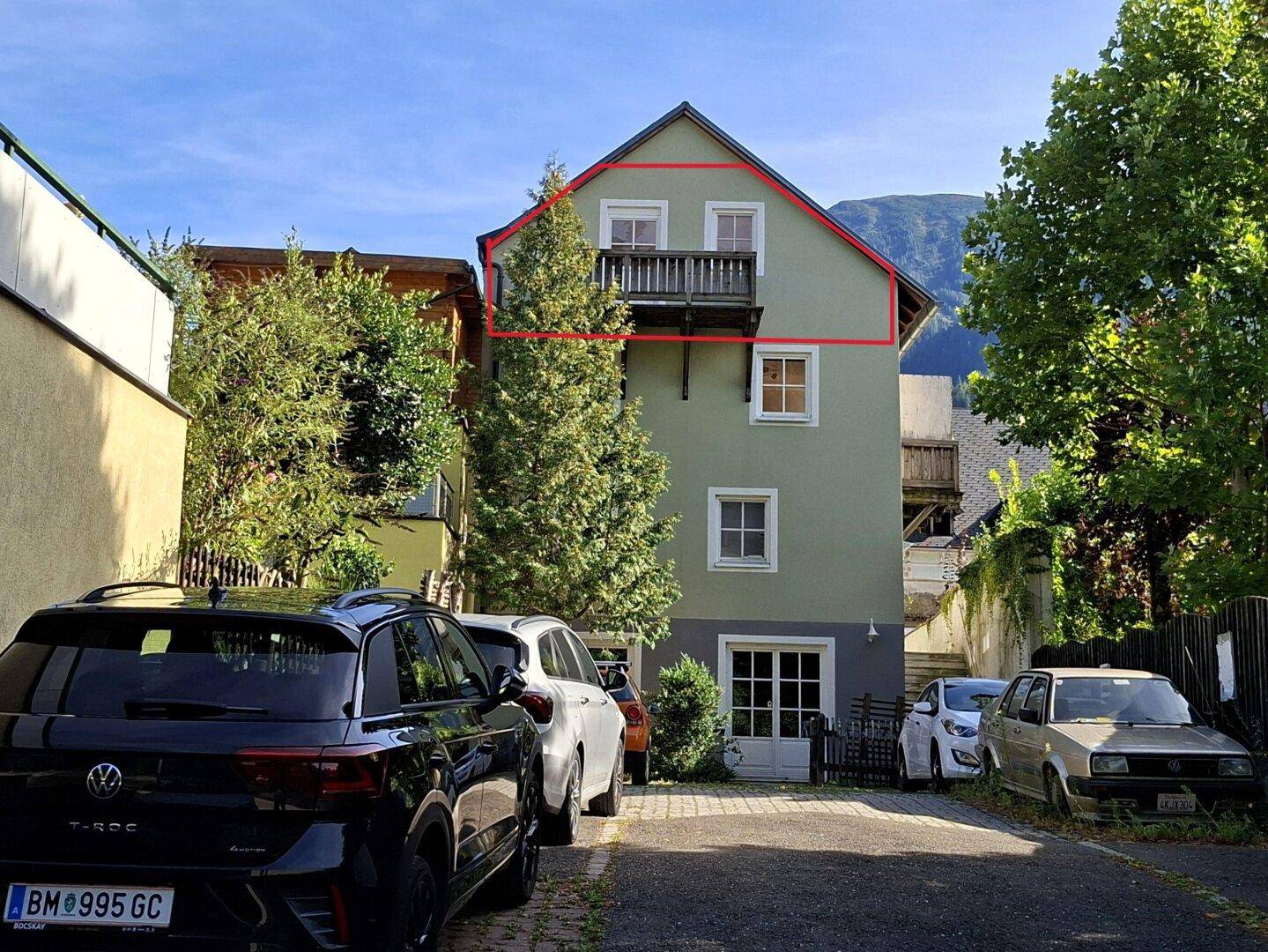 Rückansicht eines Wohnhauses mit Balkon und parkenden Autos im Vordergrund.