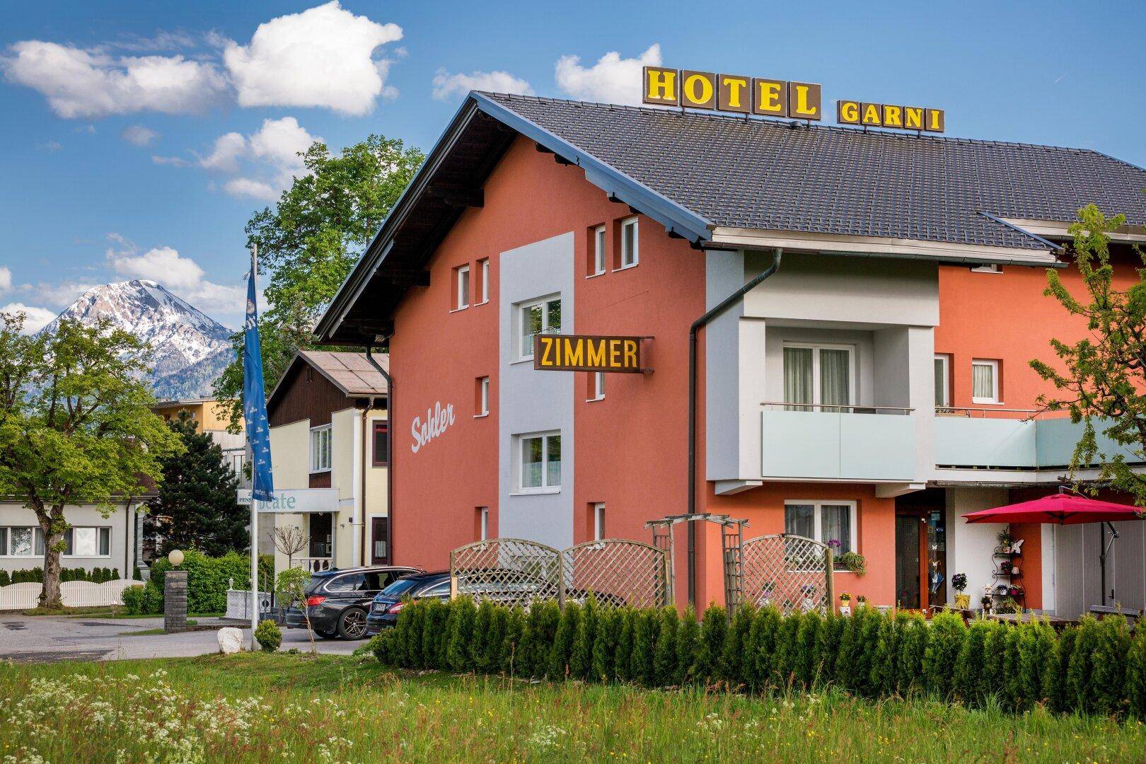 Die Fassade des Hotels mit einem roten Sonnenschirm und gepflegter Hecke vor einer Bergkulisse.