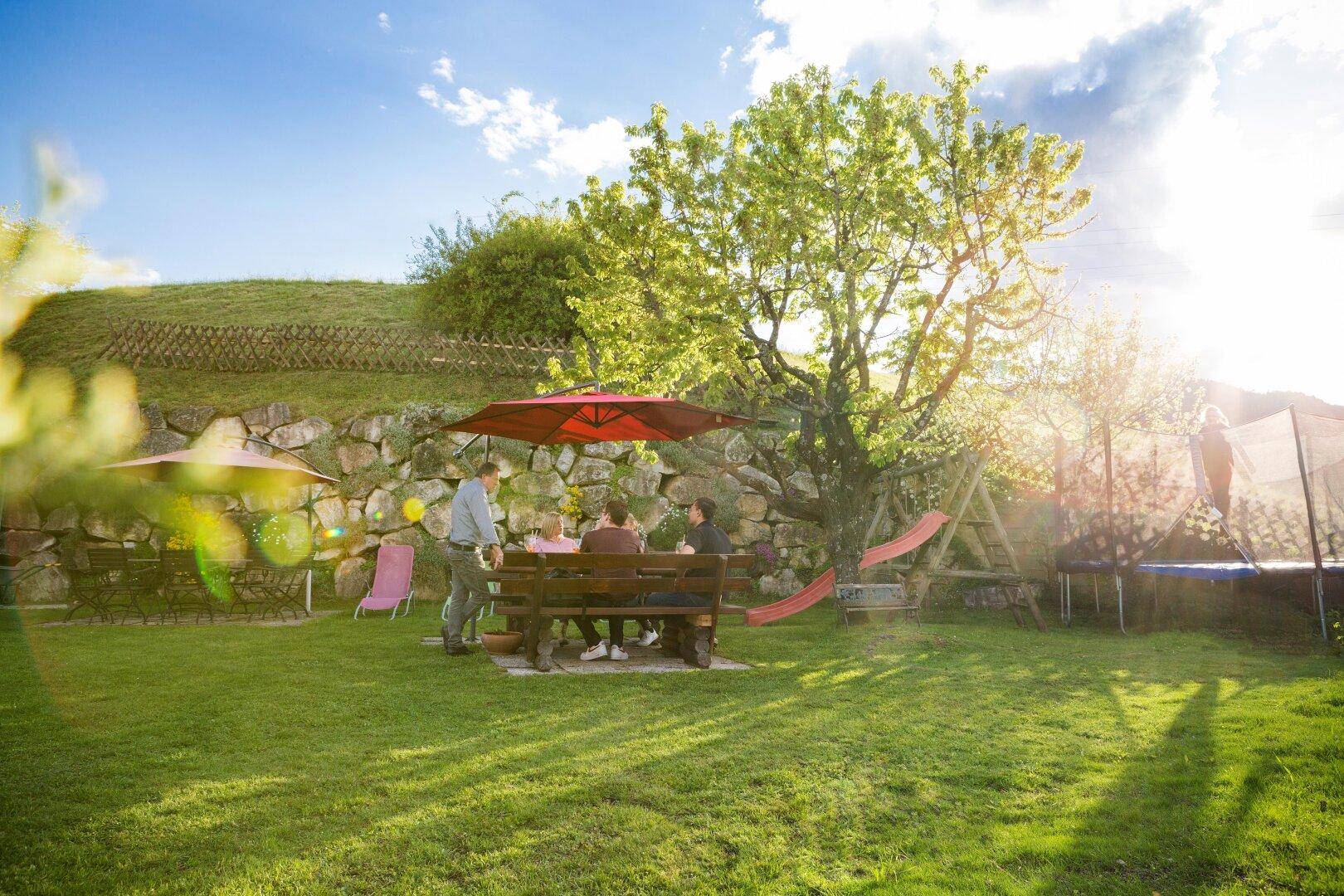 Gäste entspannen sich an einem Holztisch im sonnigen Gartenbereich mit Spielgeräten und Trampolin.