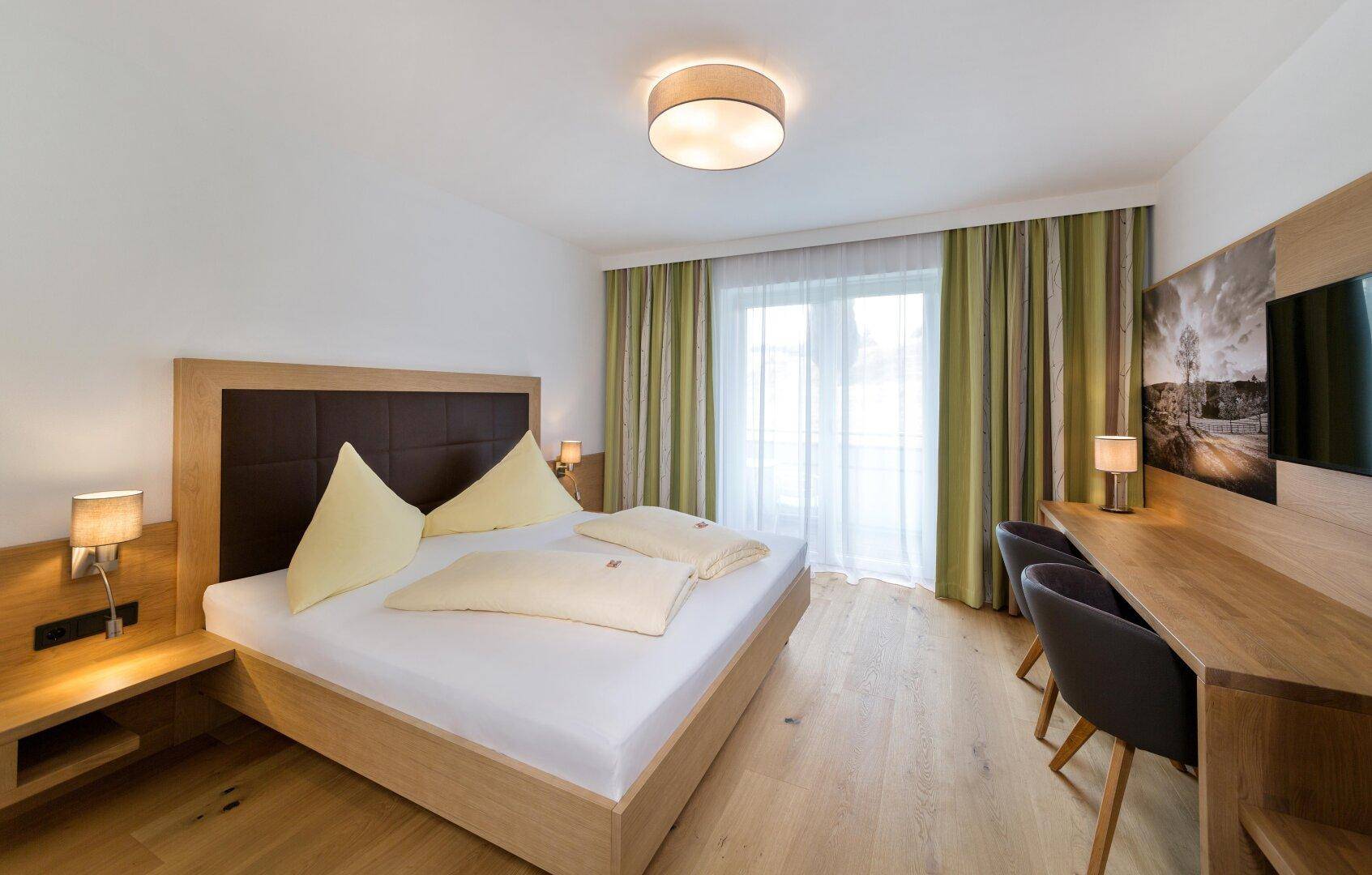 Helles Schlafzimmer mit Doppelbett, Holzboden und einem Schreibtischbereich neben dem Fenster.