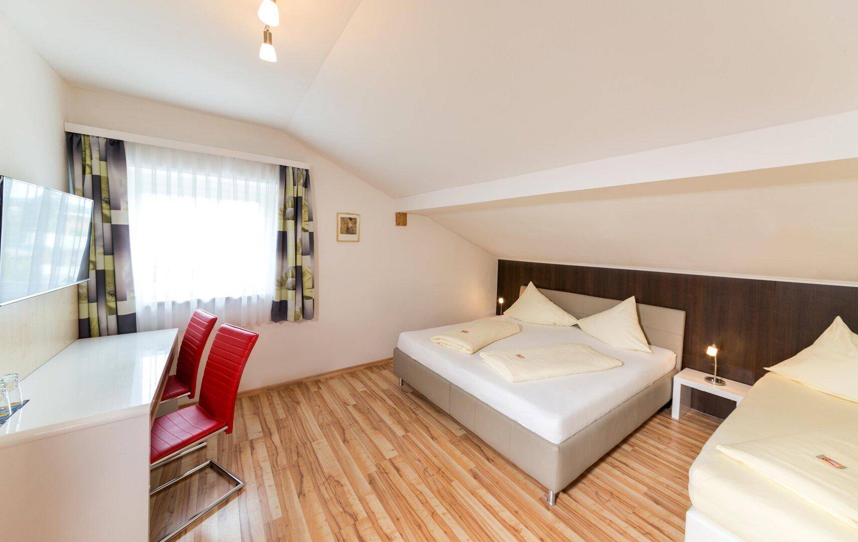 Helles Schlafzimmer im Dachgeschoss mit Doppelbett, Holzboden und rotem Schreibtischstuhl.