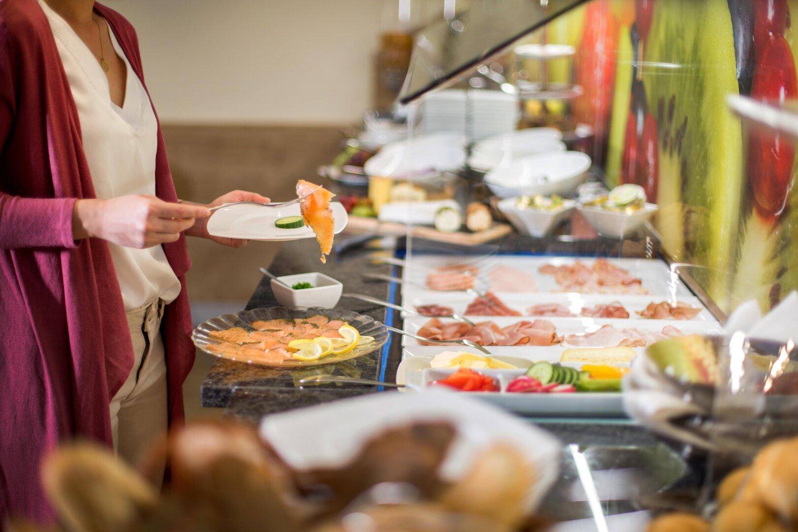 Eine Person bedient sich am vielfältigen Frühstücksbuffet mit Lachs, Aufschnitt und frischem Gemüse.