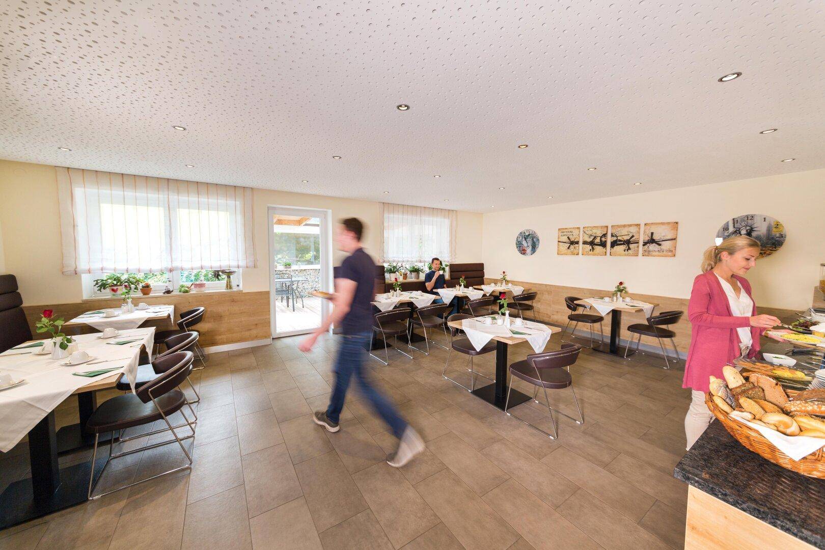 Heller Frühstücksraum mit Gästen, die sich am Buffet bedienen, und Blick auf eine Außenterrasse.