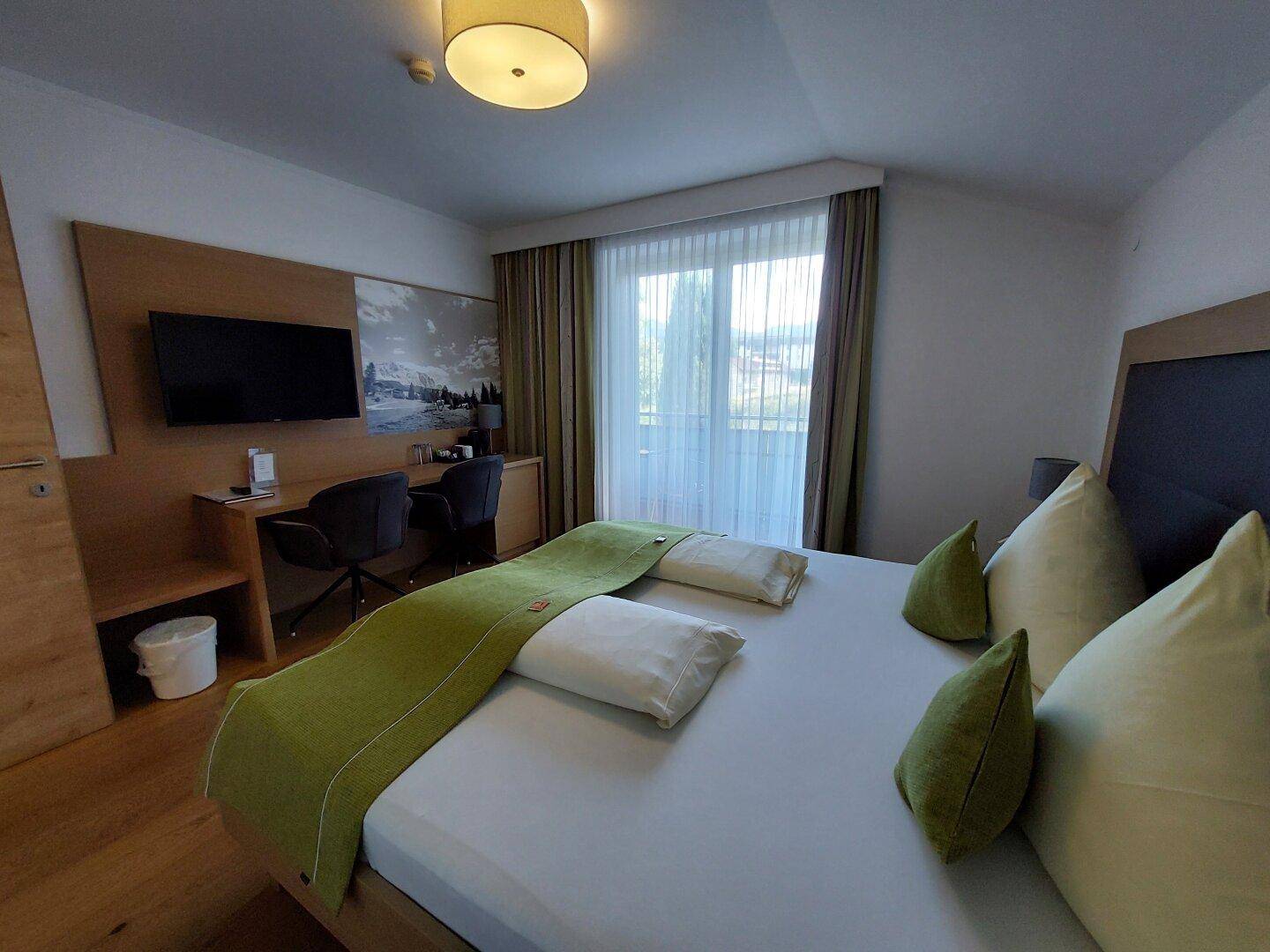 Geräumiges Schlafzimmer mit Doppelbett, Holzboden, Schreibtisch und großem Fenster mit Vorhängen.
