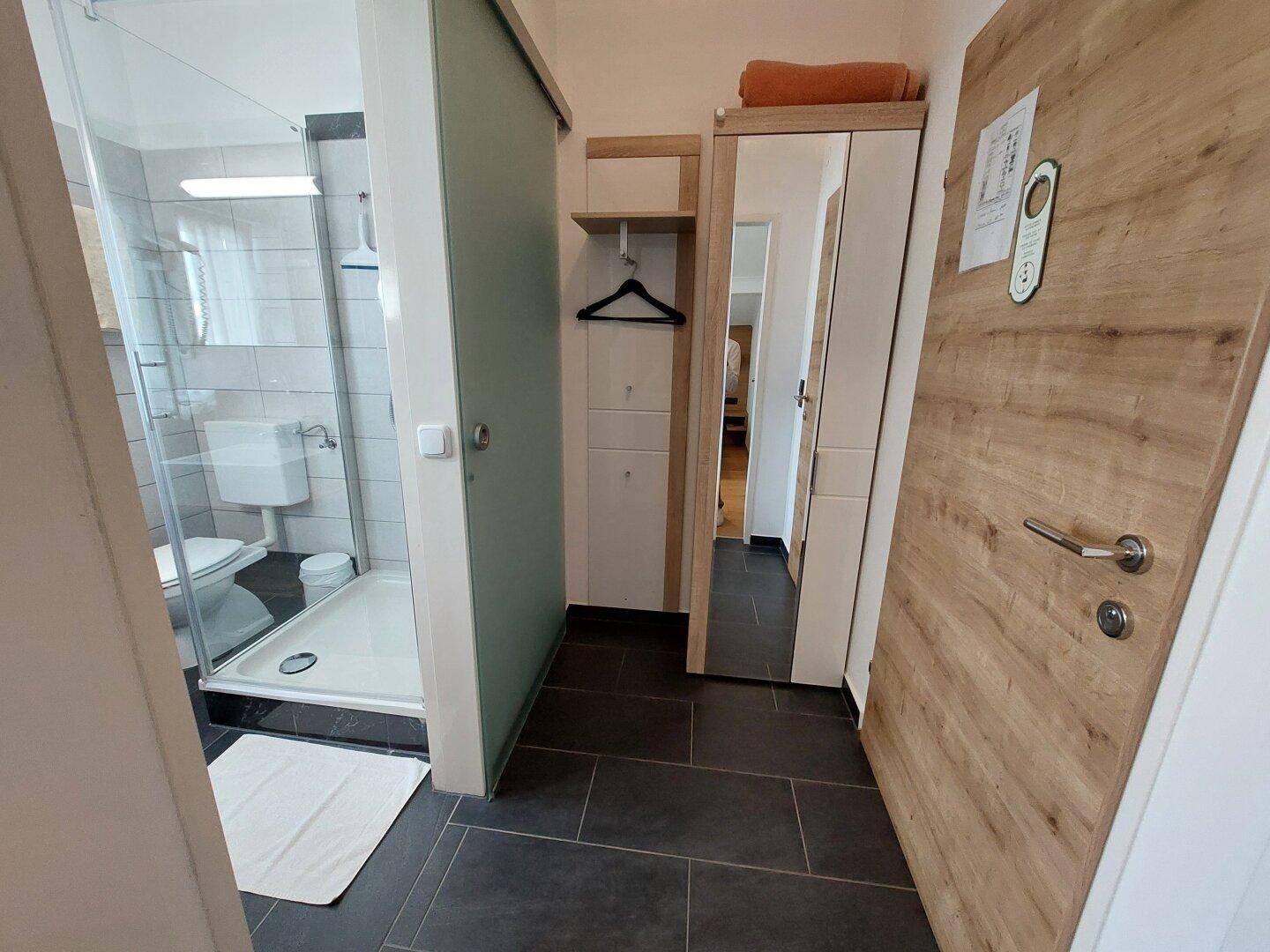 Badezimmer mit Duschkabine und Blick auf einen Flur mit Kleiderschrank und Holztür.