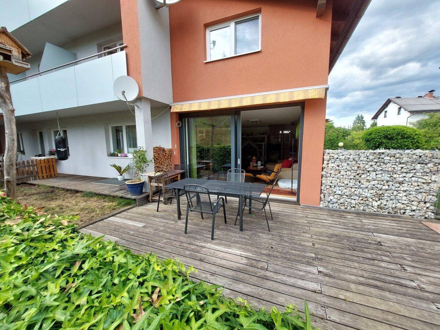 Holzterrasse mit Esstisch und Stühlen vor dem Gebäude, umgeben von Grünflächen.