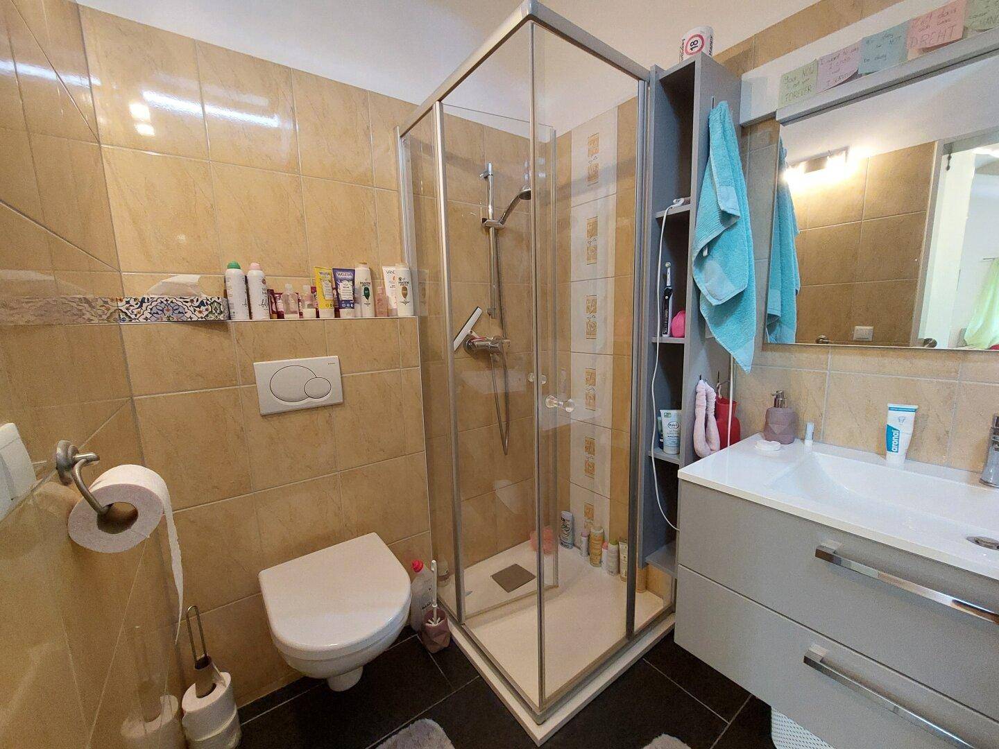 Badezimmer mit beigen Fliesen, Duschkabine, WC und modernem Waschtisch mit Spiegel.