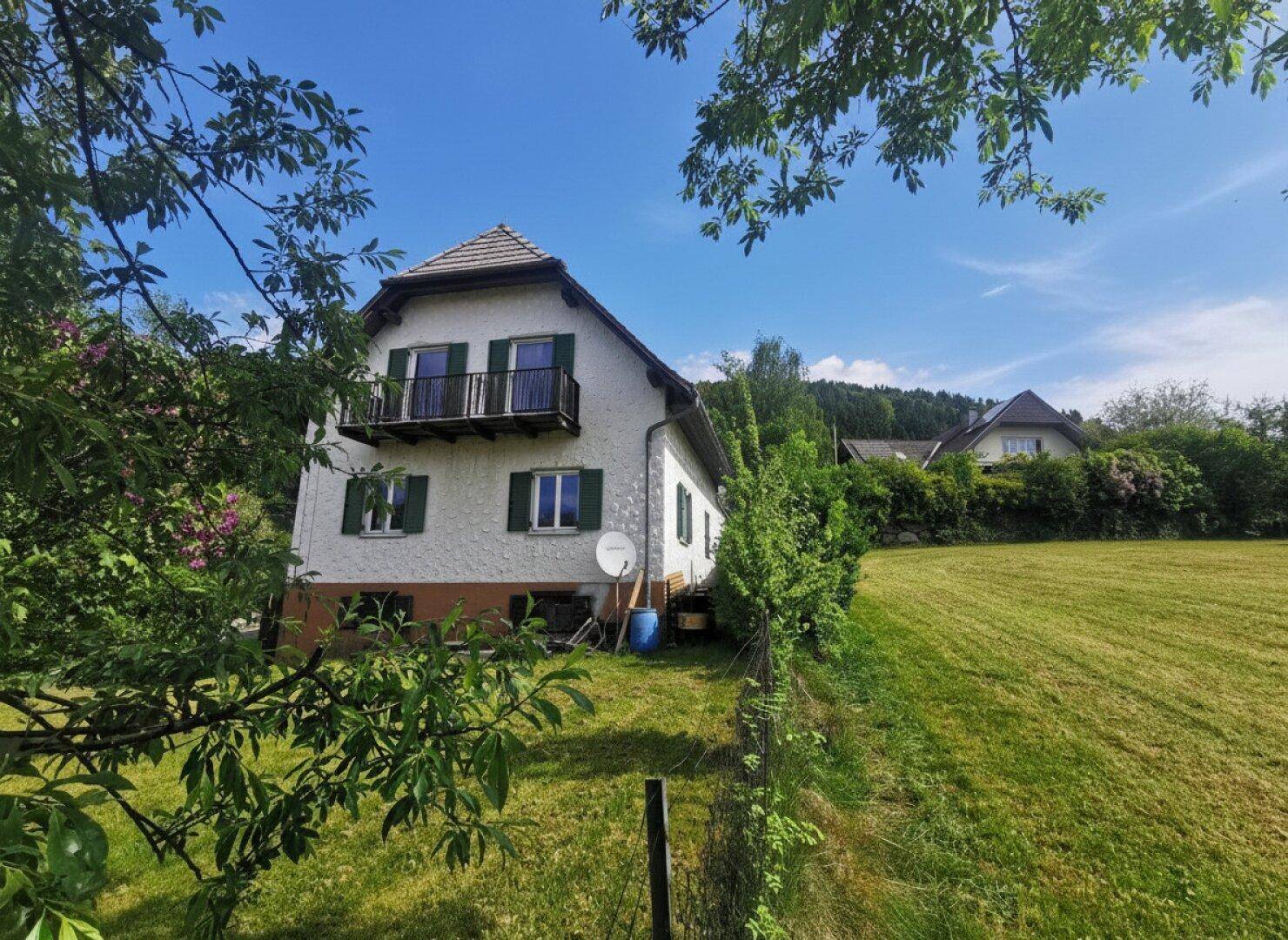 Freistehendes Haus mit Balkon und grüner Umgebung, umgeben von Bäumen und Wiesen unter blauem Himmel.