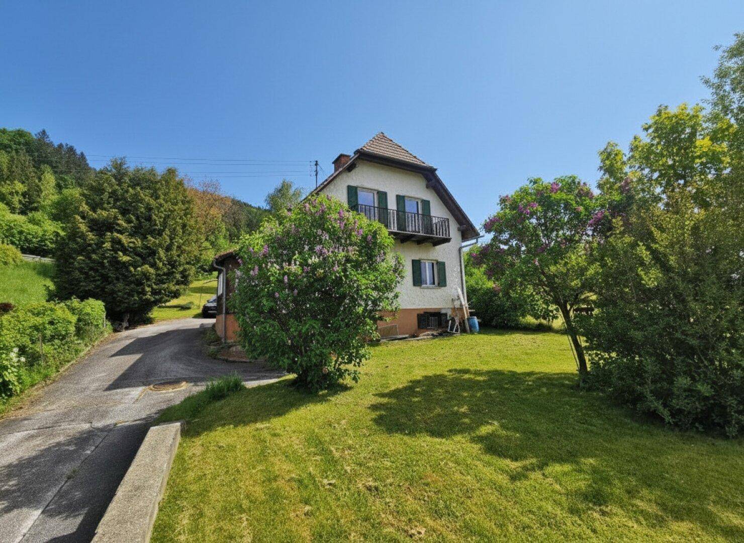 Einfamilienhaus mit großem Garten und Zufahrt, umgeben von blühenden Büschen und Bäumen im Sonnenschein.