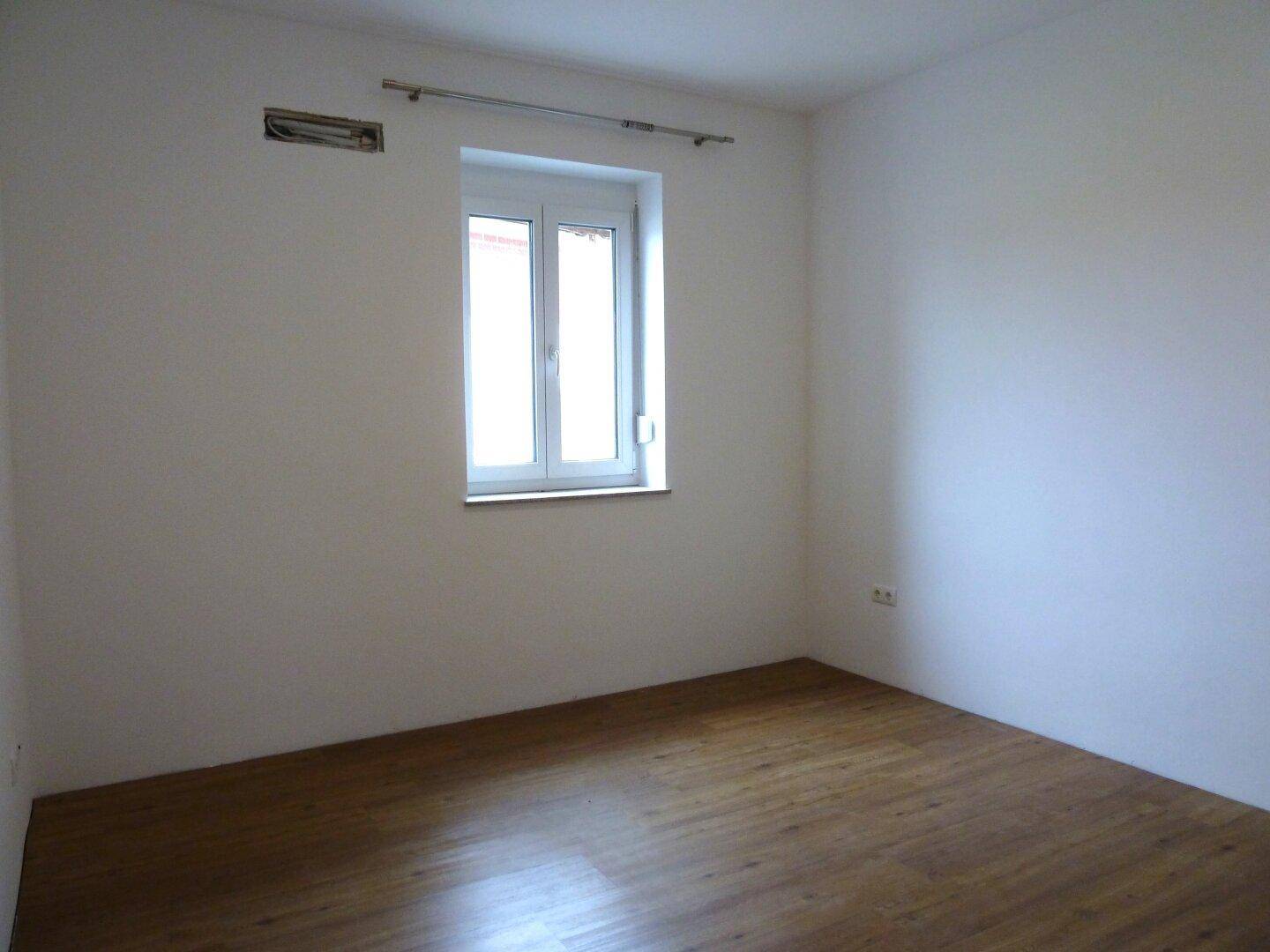 Helles, leeres Zimmer mit Holzfußboden und einem Fenster, bereit zur individuellen Gestaltung.