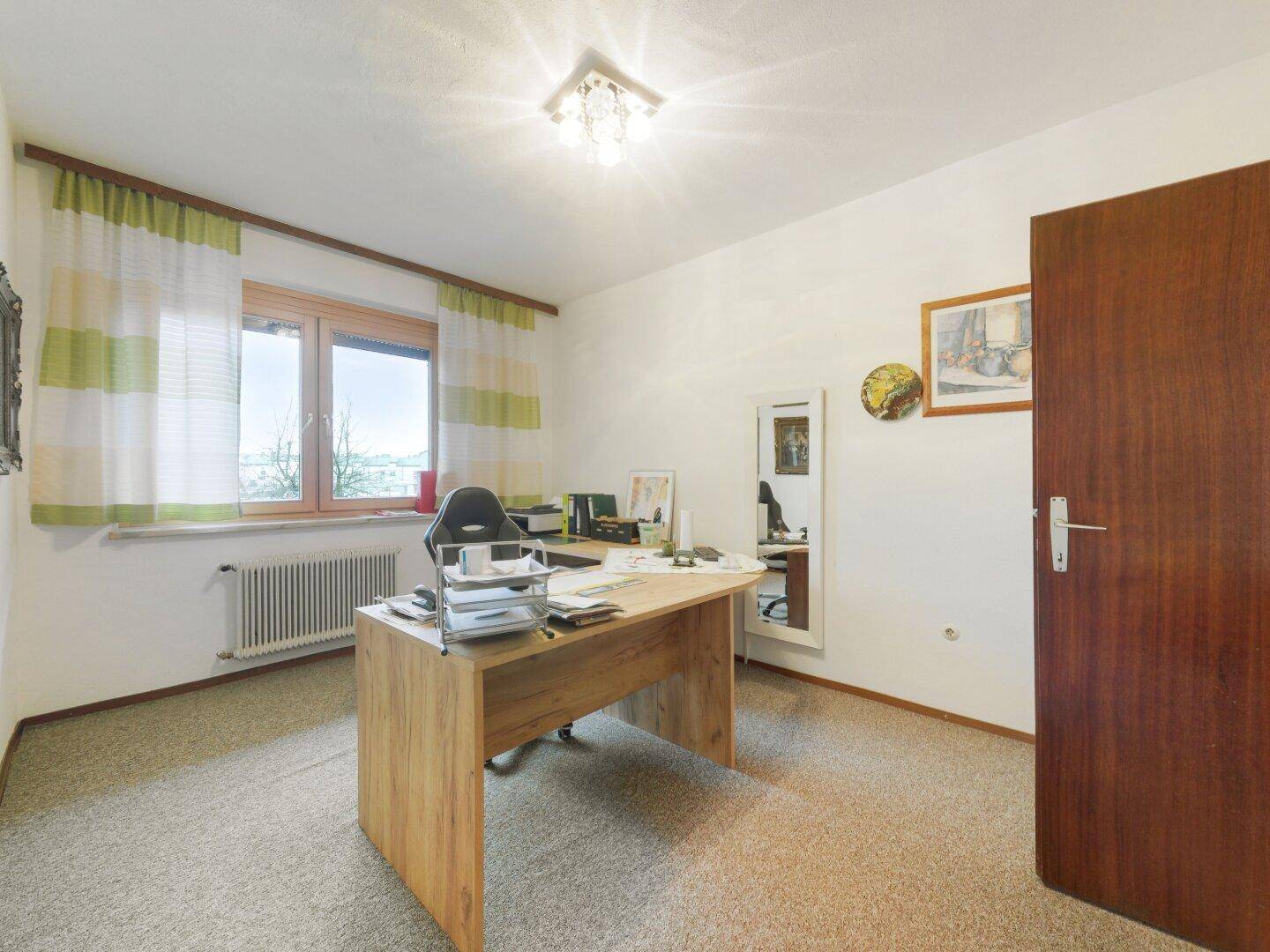 Helles Büro mit Teppichboden, Holzschreibtisch und Blick ins Grüne durch das Fenster.