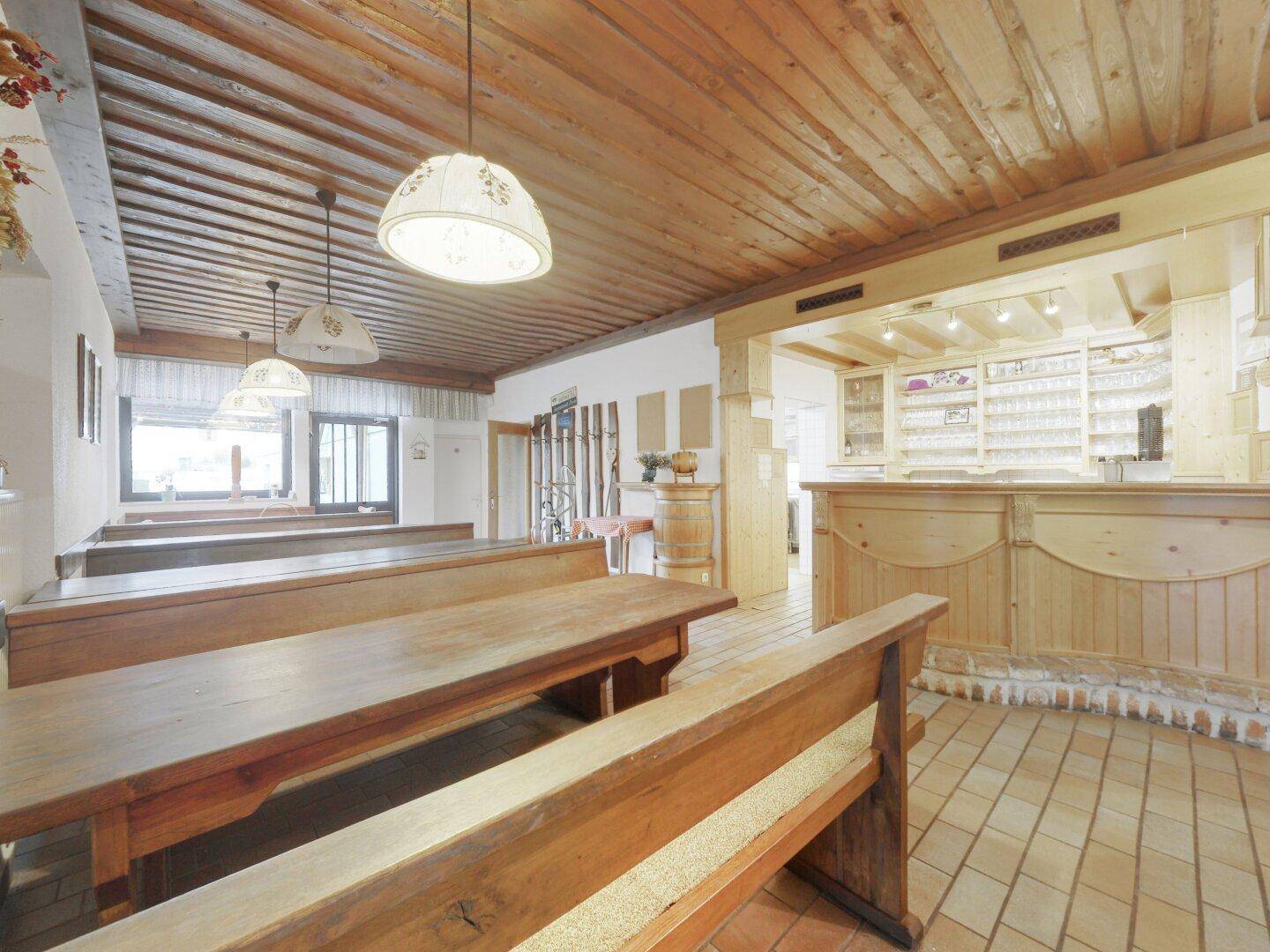 Rustikaler Speisesaal mit langen Holzbänken, gefliestem Boden und einer Theke im Hintergrund.