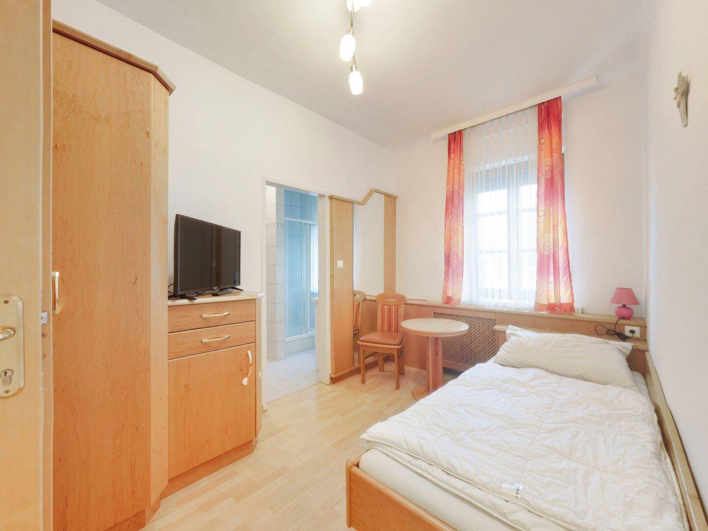 Kleines Schlafzimmer mit Einzelbett, Holzschrank und Zugang zu einem angrenzenden Badezimmer.