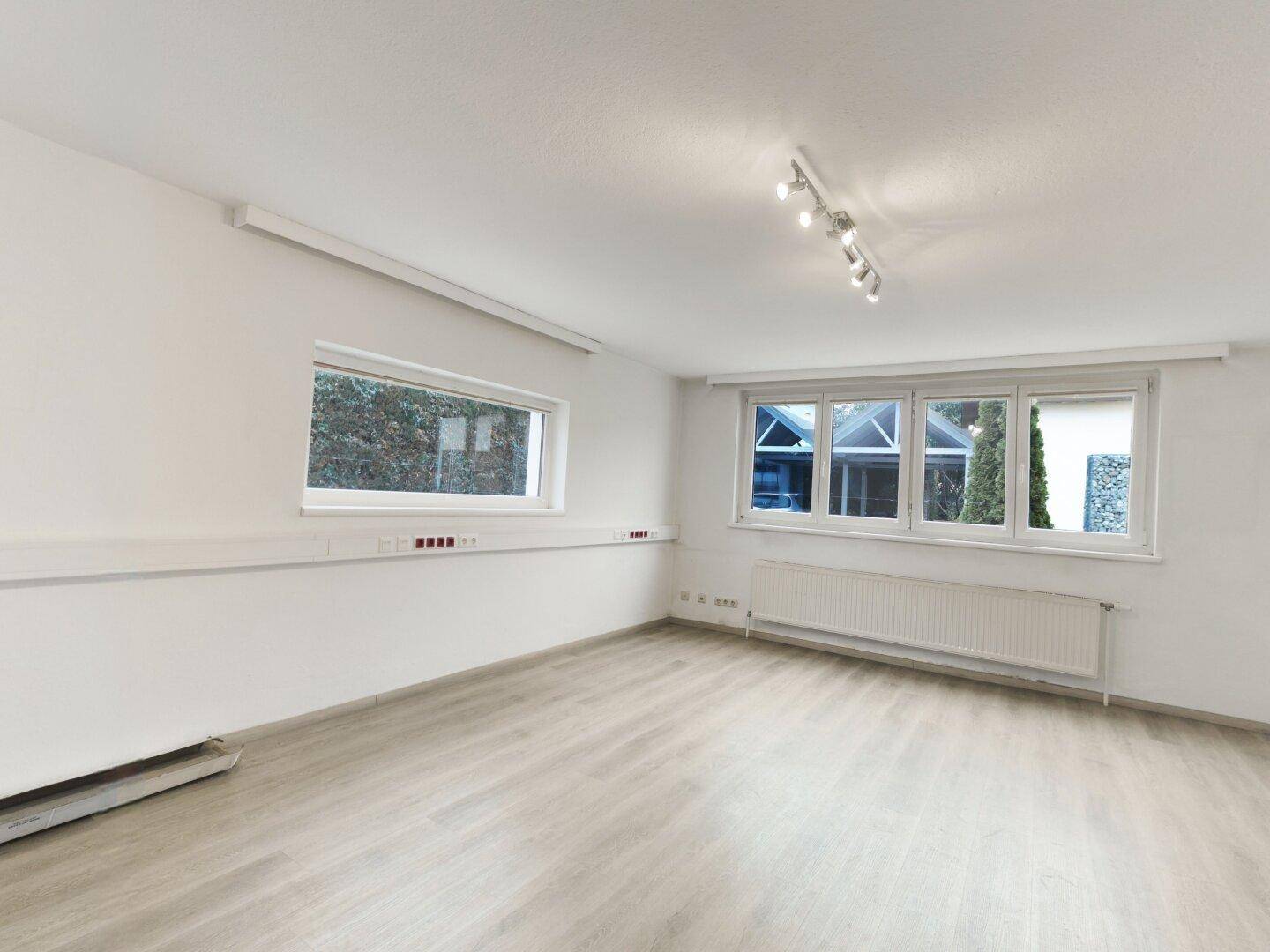 Geräumiger, heller Raum mit mehreren Fenstern, Heizkörpern und modernem Laminatboden, bereit zur Einrichtung.