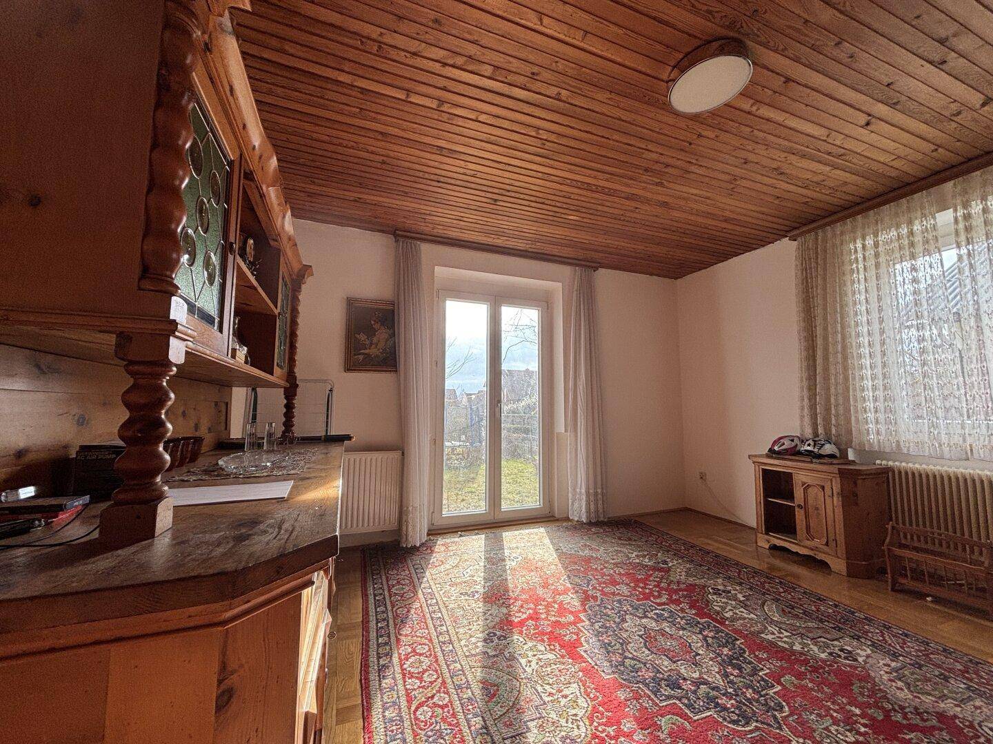 Wohnzimmer mit Holzbalkendecke, Teppich und Zugang zu einem sonnigen Außenbereich.