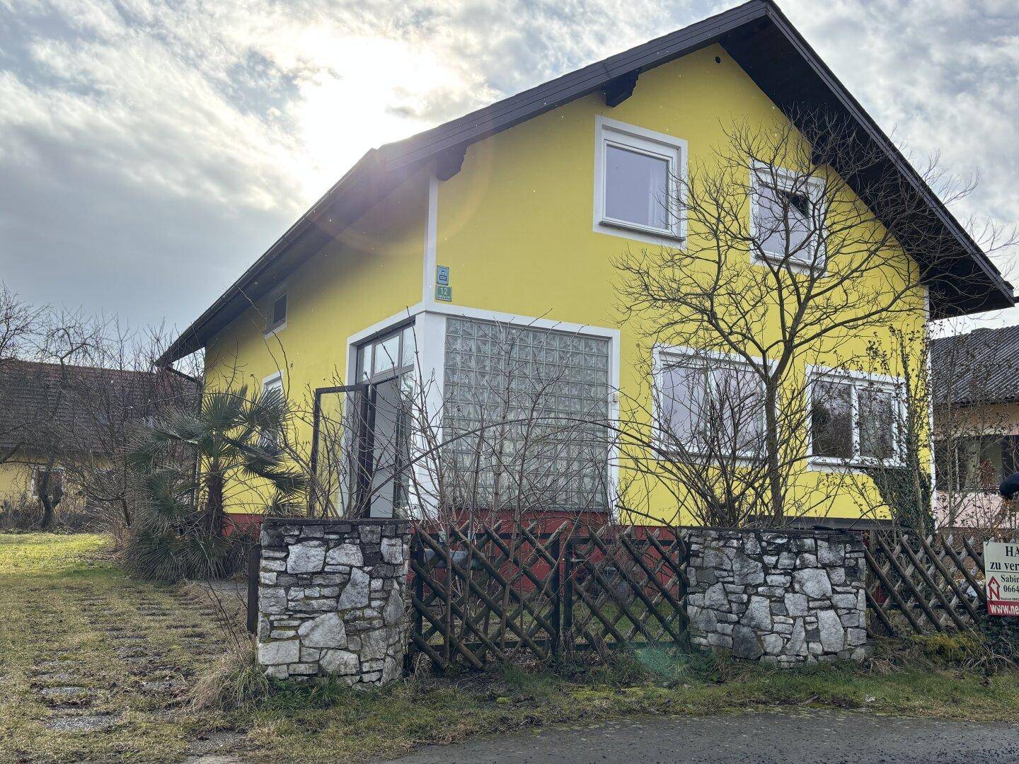 Gelbes Einfamilienhaus mit Steinfassade und Holzzaun an einem sonnigen Tag.