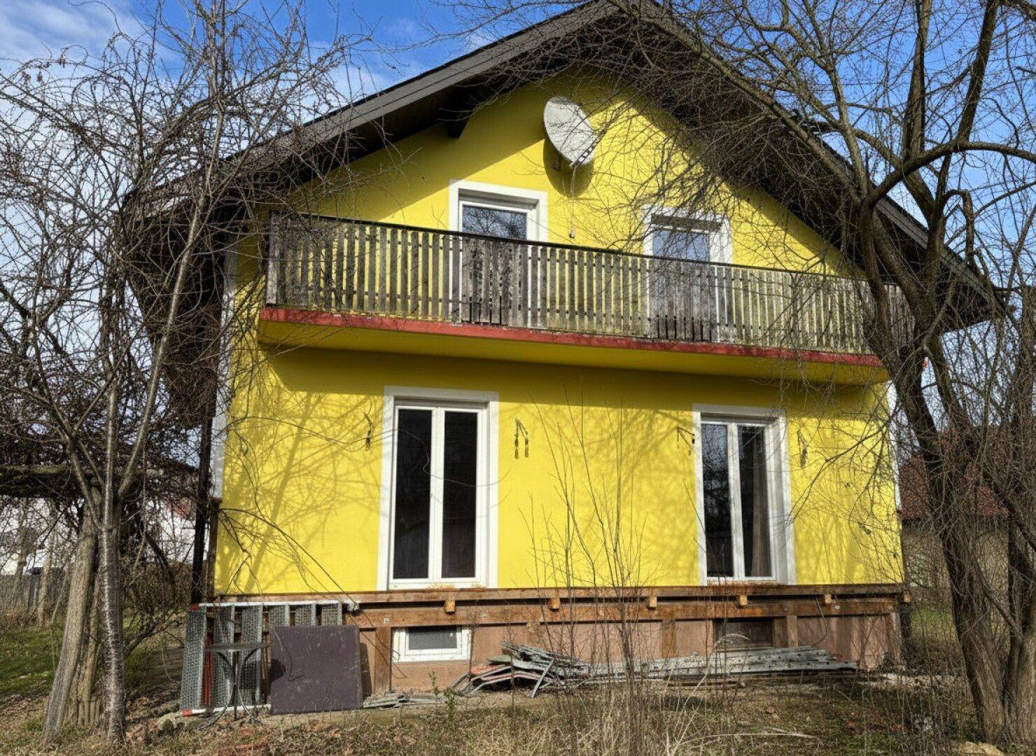 Gelbes Einfamilienhaus mit Balkon und großen Fenstern, umgeben von Bäumen.
