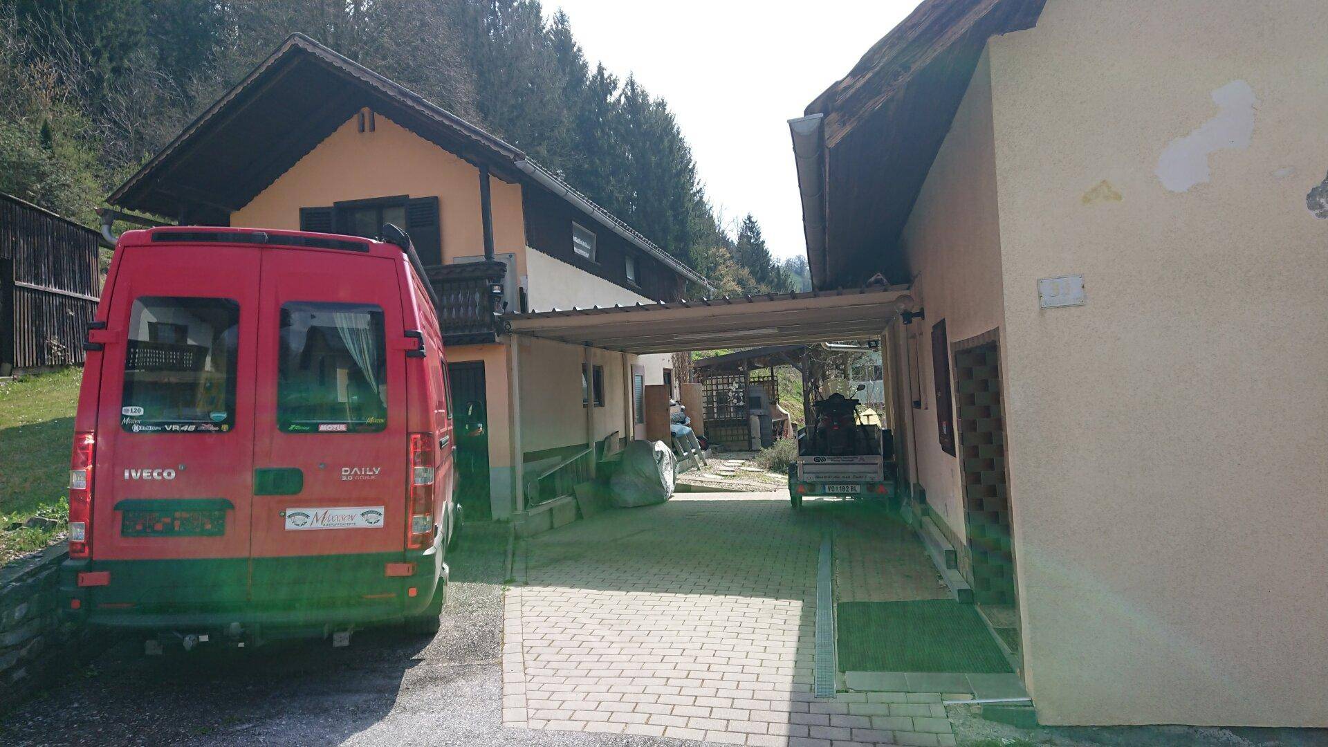 Einfahrt mit gepflastertem Bereich, Carport und einem roten Lieferwagen vor dem Haus.