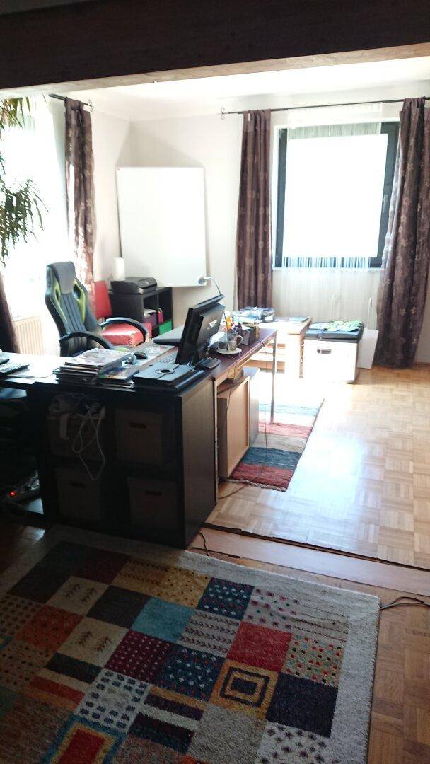 Helles Arbeitszimmer mit Parkettboden, großem Fenster und einem bunten Teppich.