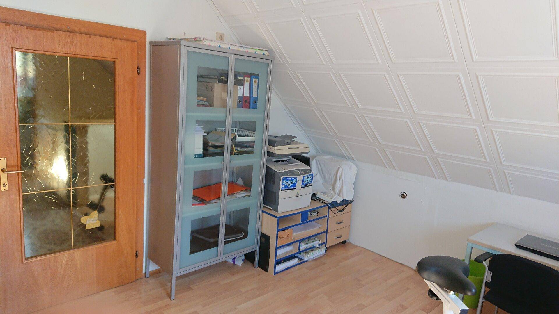 Dachzimmer mit schräger Decke, Laminatboden und einem Schrank für Bürobedarf.