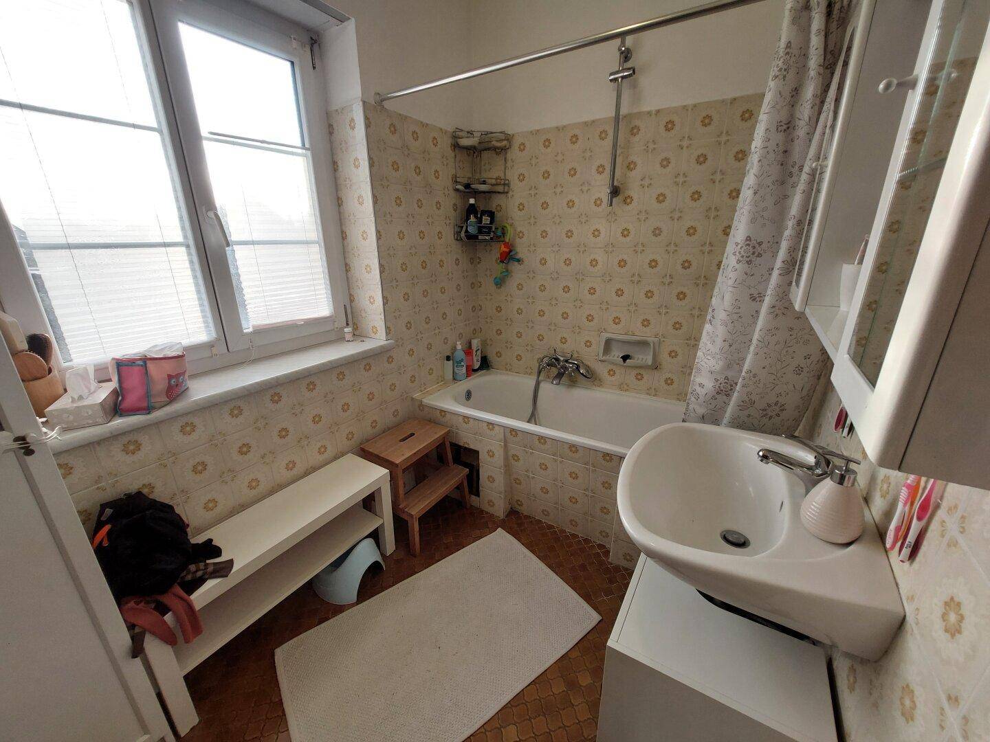 Badezimmer mit Badewanne, Waschbecken und Fenster, gefliest im 80er-Jahre-Stil.
