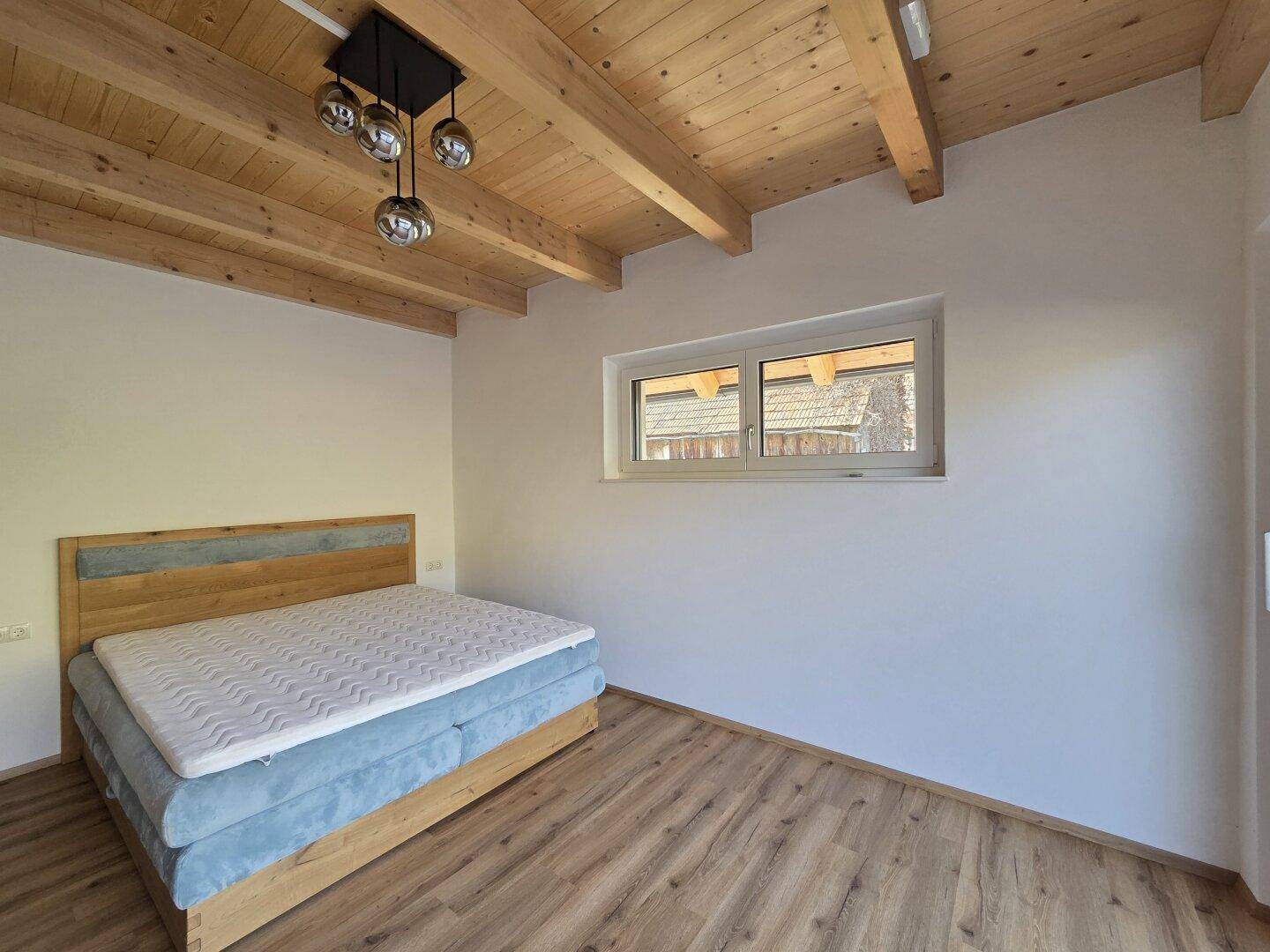Schlafzimmer mit Holzbalkendecke, Laminatboden und einem Fenster mit Blick ins Grüne.