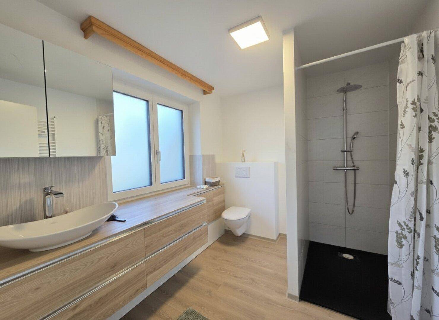 Helles, modernes Badezimmer mit Fenster, Holzwaschtisch, wandhängendem WC und Duschbereich.