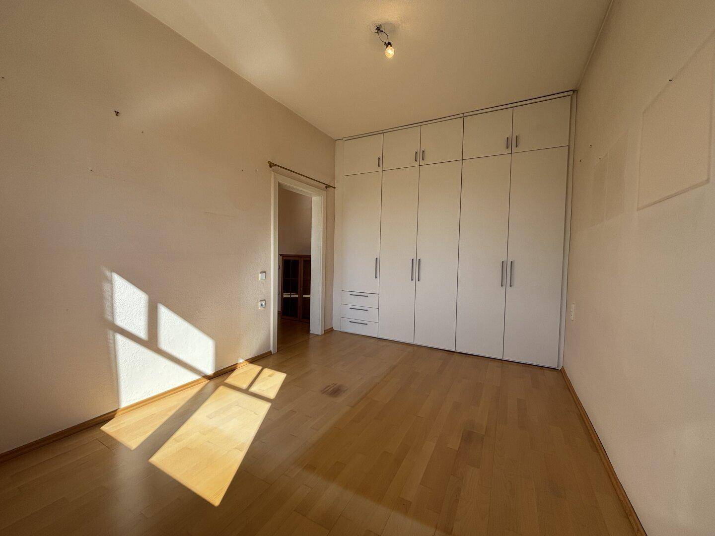 Geräumiger Raum mit Einbauschränken, Parkettboden und sonnigen Lichtflecken.