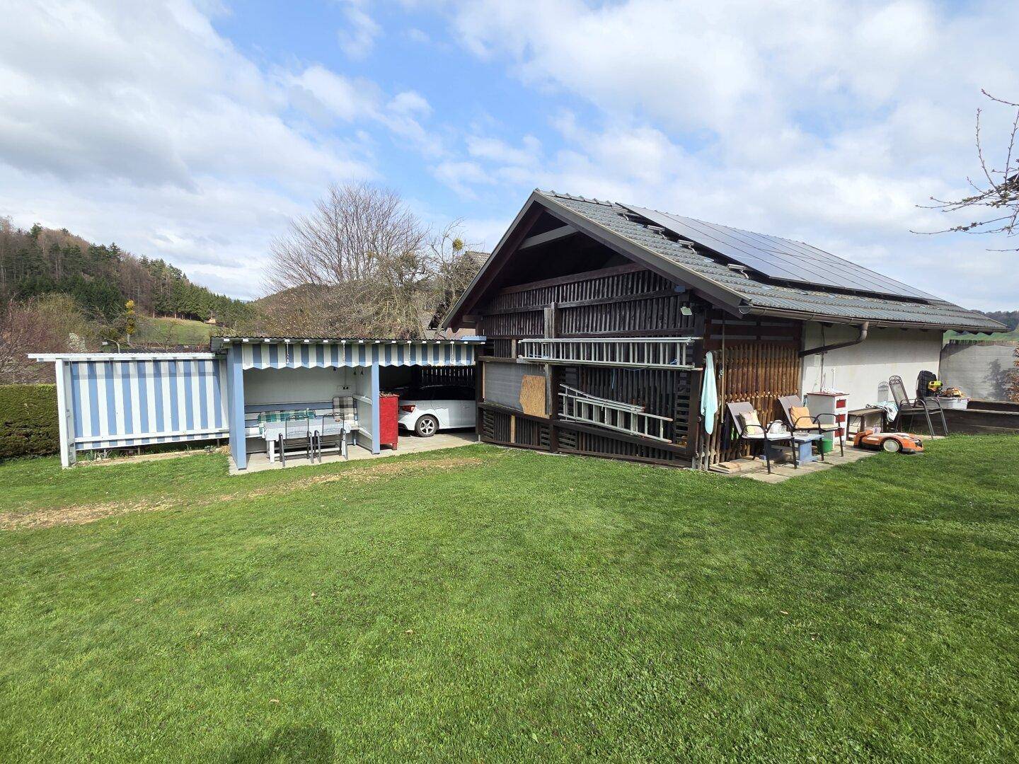Gartenbereich mit einem Holzschuppen, Solarpaneelen auf dem Dach und einem überdachten Sitzbereich.