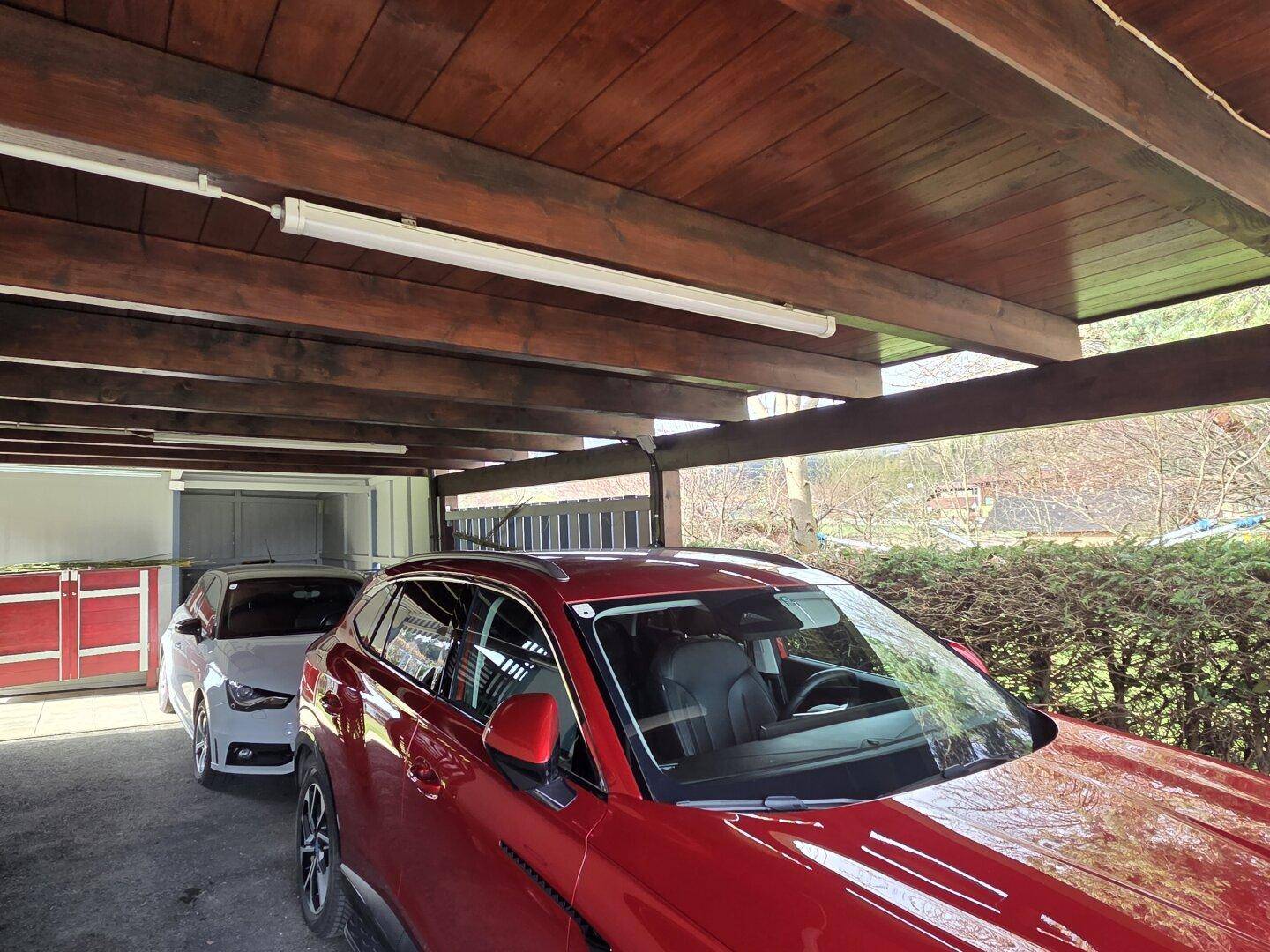 Überdachter Carport mit Holzbalken und zwei geparkten Autos, angrenzend an eine Hecke.