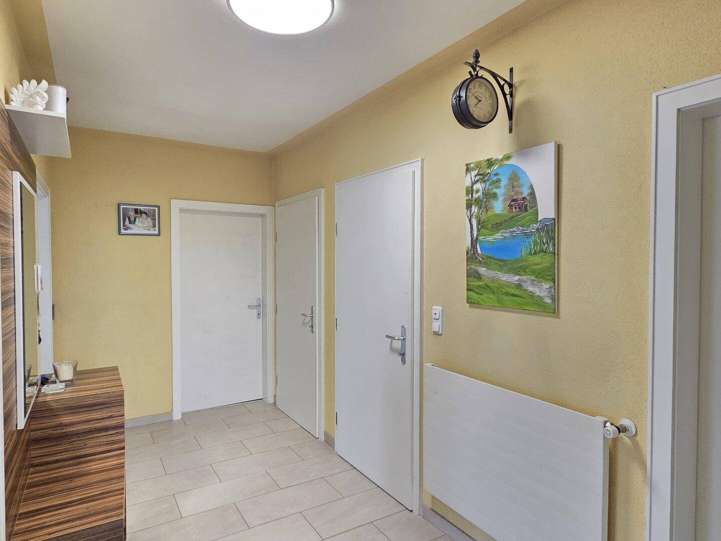 Geräumiger Flur mit hellen Fliesen, gelben Wänden und einem Heizkörper unter einem Wandbild.