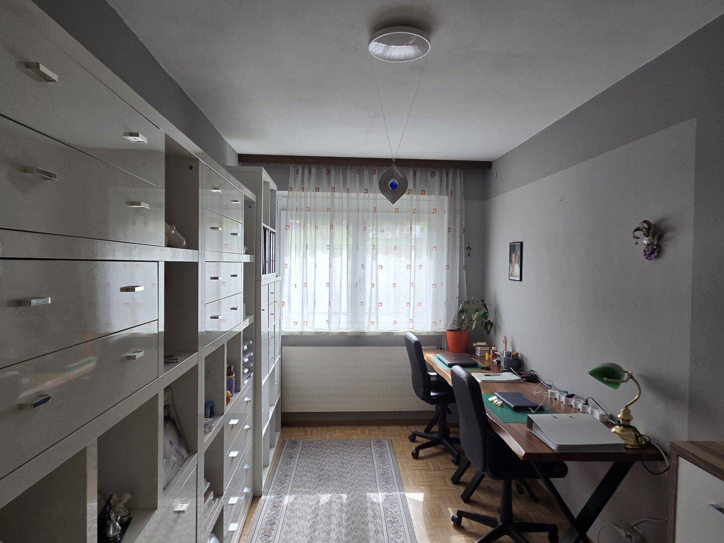Funktionales Arbeitszimmer mit Regalsystemen, zwei Schreibtischen und einem Fenster mit Vorhängen.