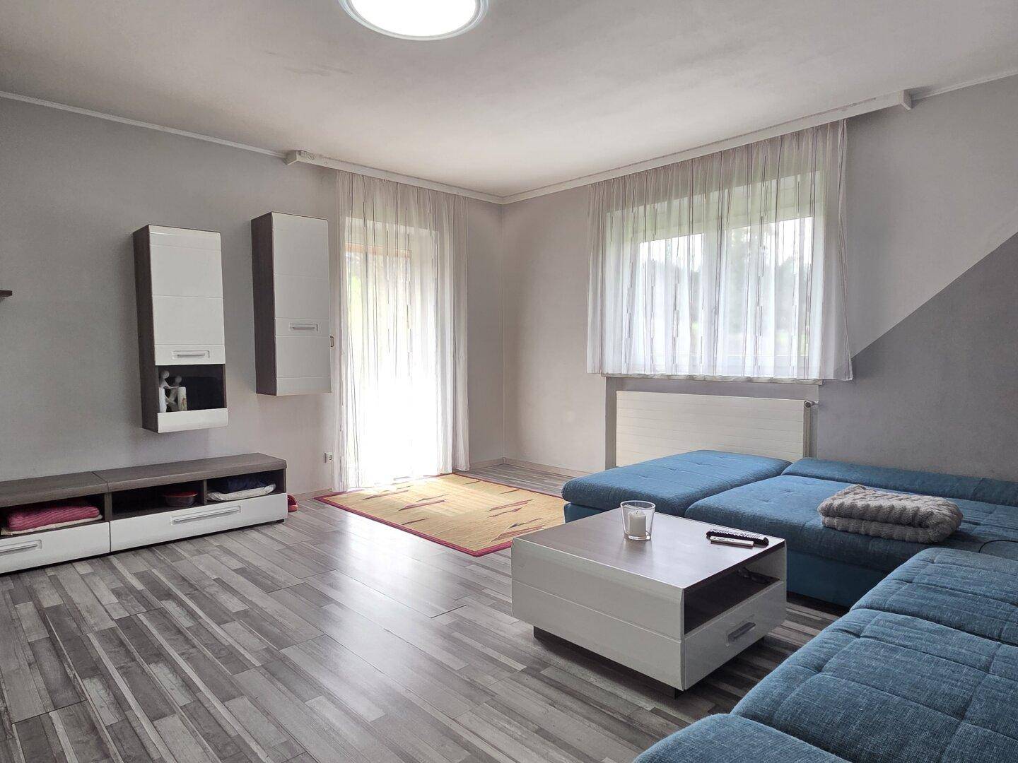 Gemütliches Wohnzimmer mit blauem Ecksofa, Laminatboden und großen Fenstern für viel Tageslicht.
