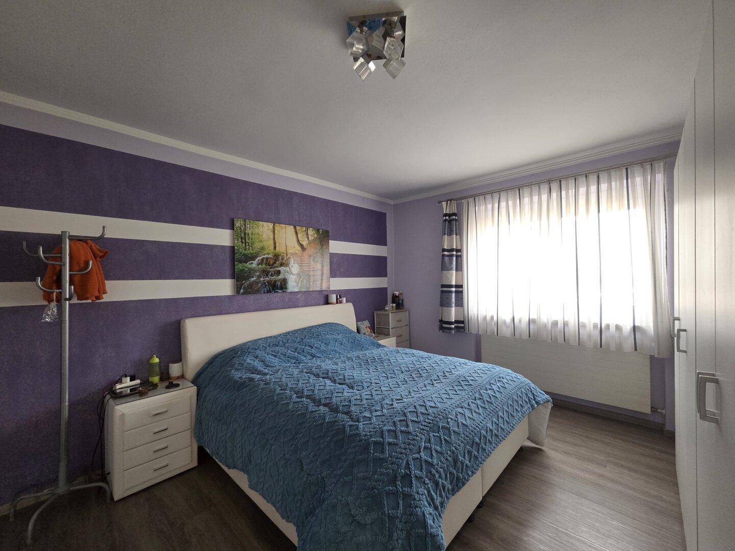 Schlafzimmer mit lila-weiß gestreiften Wänden, Doppelbett und Laminatboden, hell durch großes Fenster.