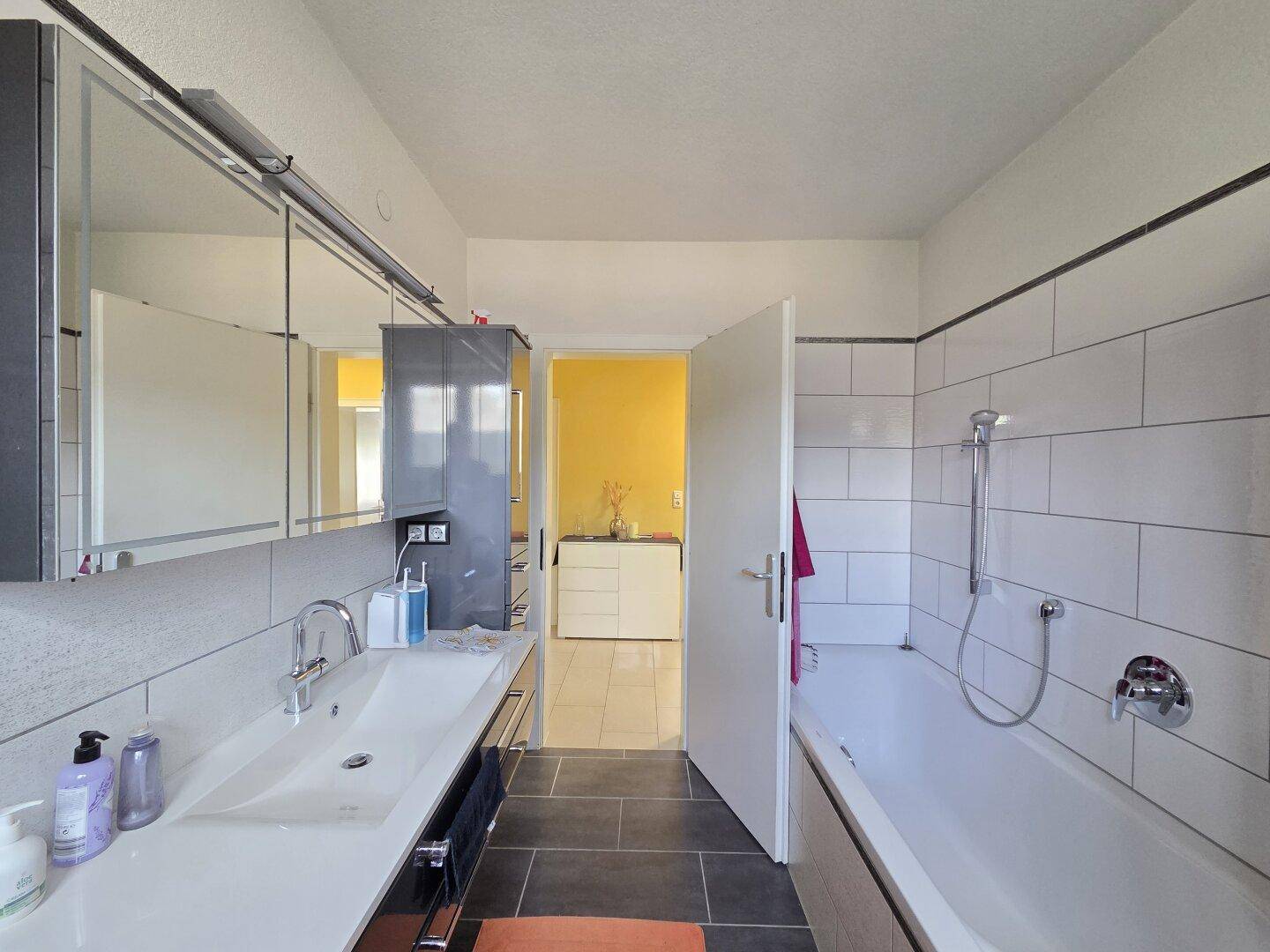 Helles Badezimmer mit Badewanne, Waschbecken und Spiegelschrank, verbunden mit dem Flur.