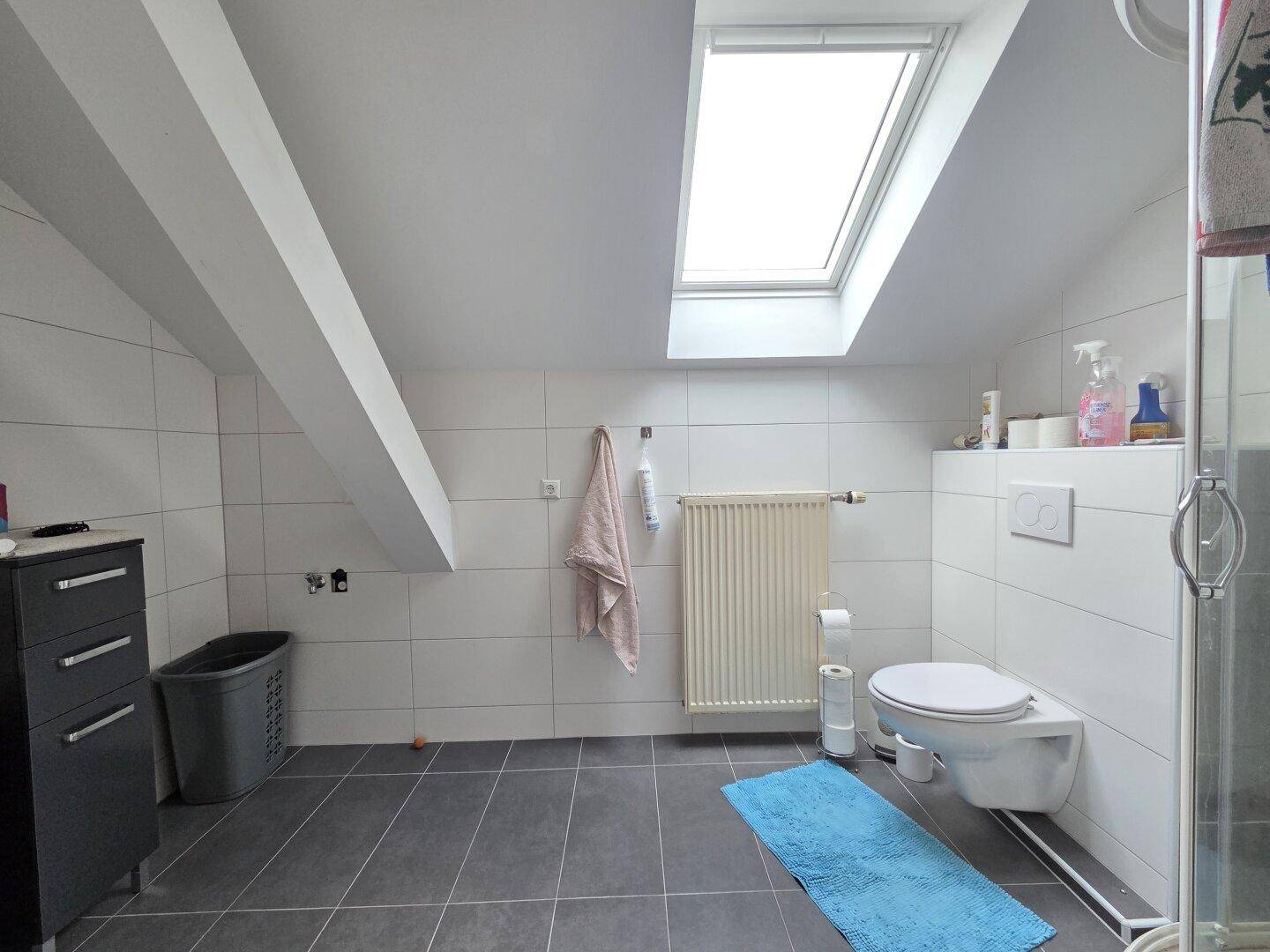 Badezimmer im Dachgeschoss mit Schrägen, weißen Fliesen, dunklem Boden und einem Dachfenster.